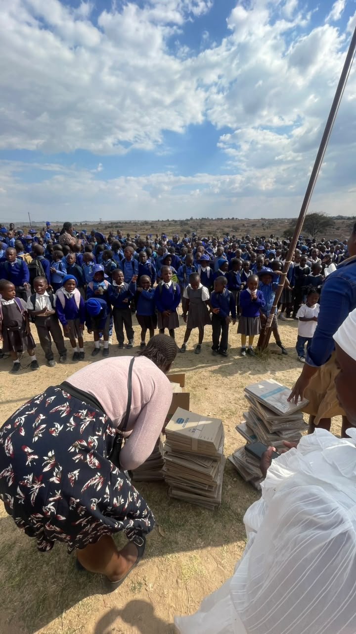 Large group of school children in uniforms gathered outdoors, with some receiving newspapers and magazines from adults.