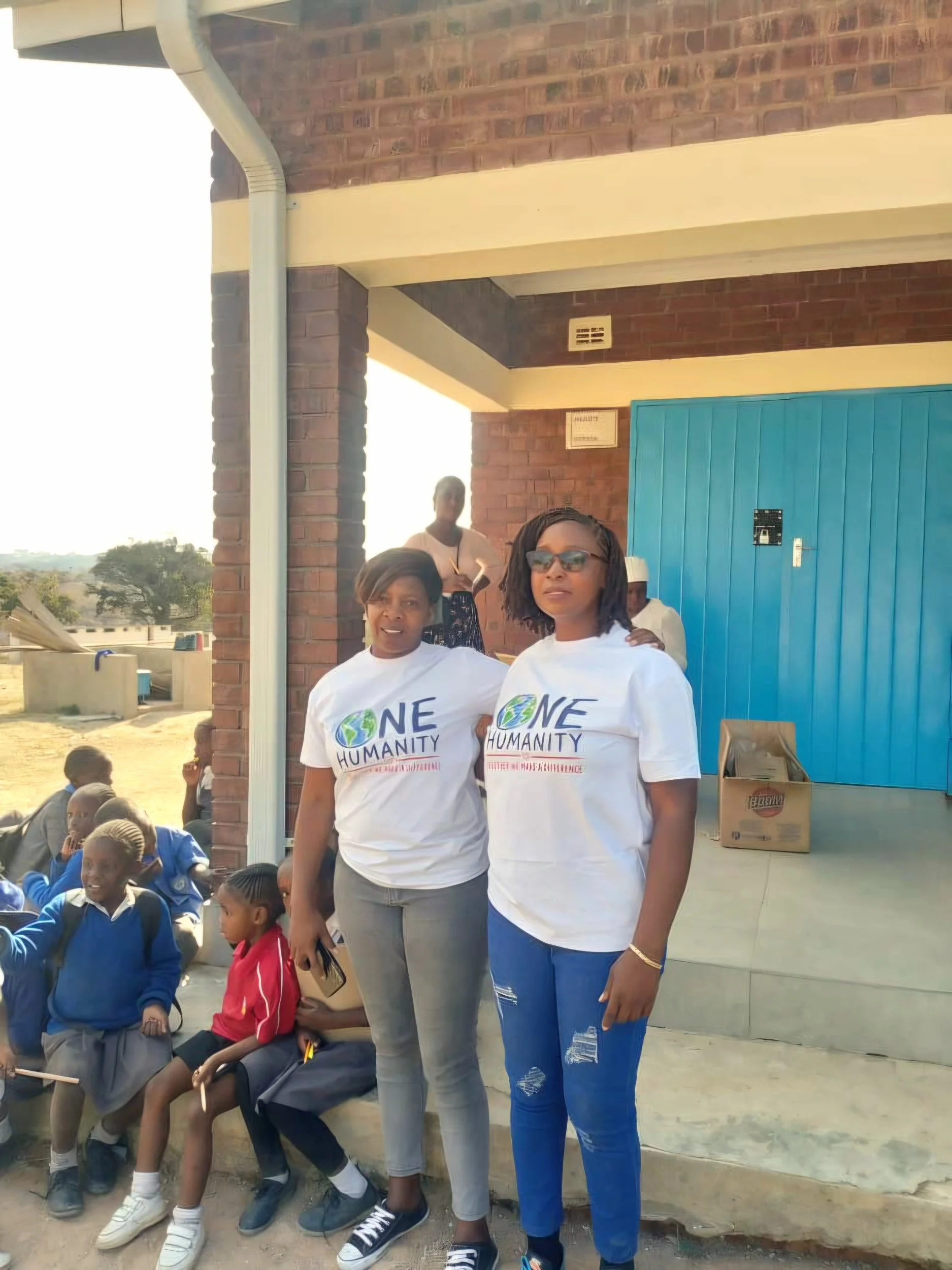 Two women standing outside in front of a brick building with children sitting nearby on the ground. Both women are wearing white t-shirts with