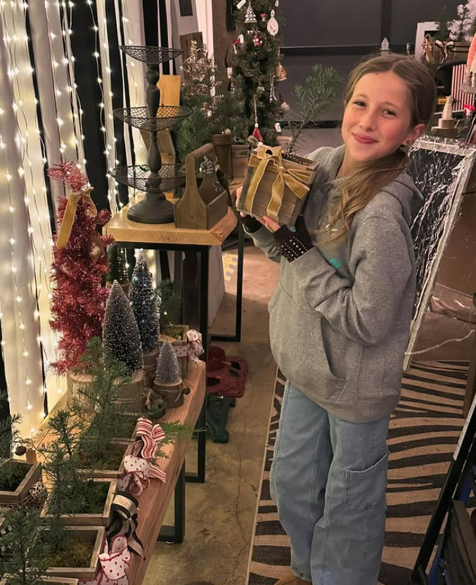 Girl holding a wrapped Christmas gift in a holiday-themed store, surrounded by small decorated Christmas trees and festive decor.