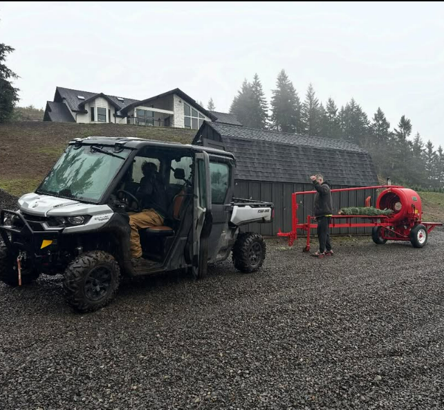A farm scene with a side-by-side utility vehicle and a man attaching a red hay baler, with a house and trees in the background on a gravel driveway.