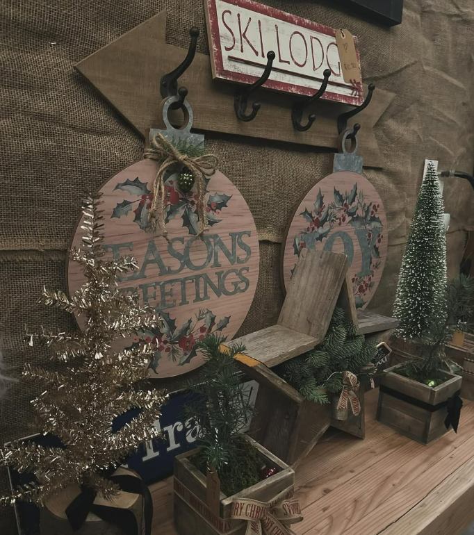 Decorative Christmas scene with signs reading 'Seasons Greetings' and 'Joy,' small artificial Christmas trees, and wooden accents in a rustic setting.