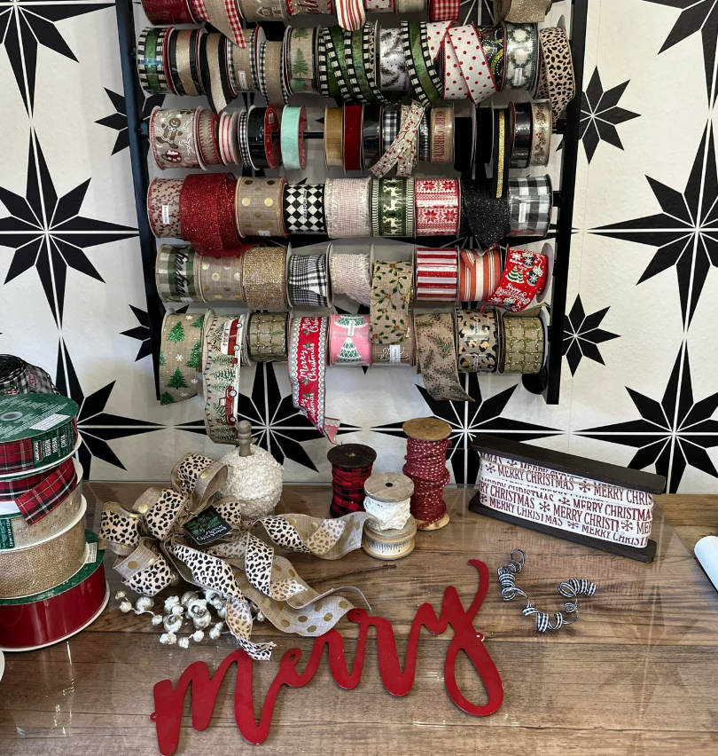 Display of Christmas ribbon rolls, a festive 'merry' sign, and holiday decorative items on a wooden table, with a background of ribbon organized on wall-mounted shelves.