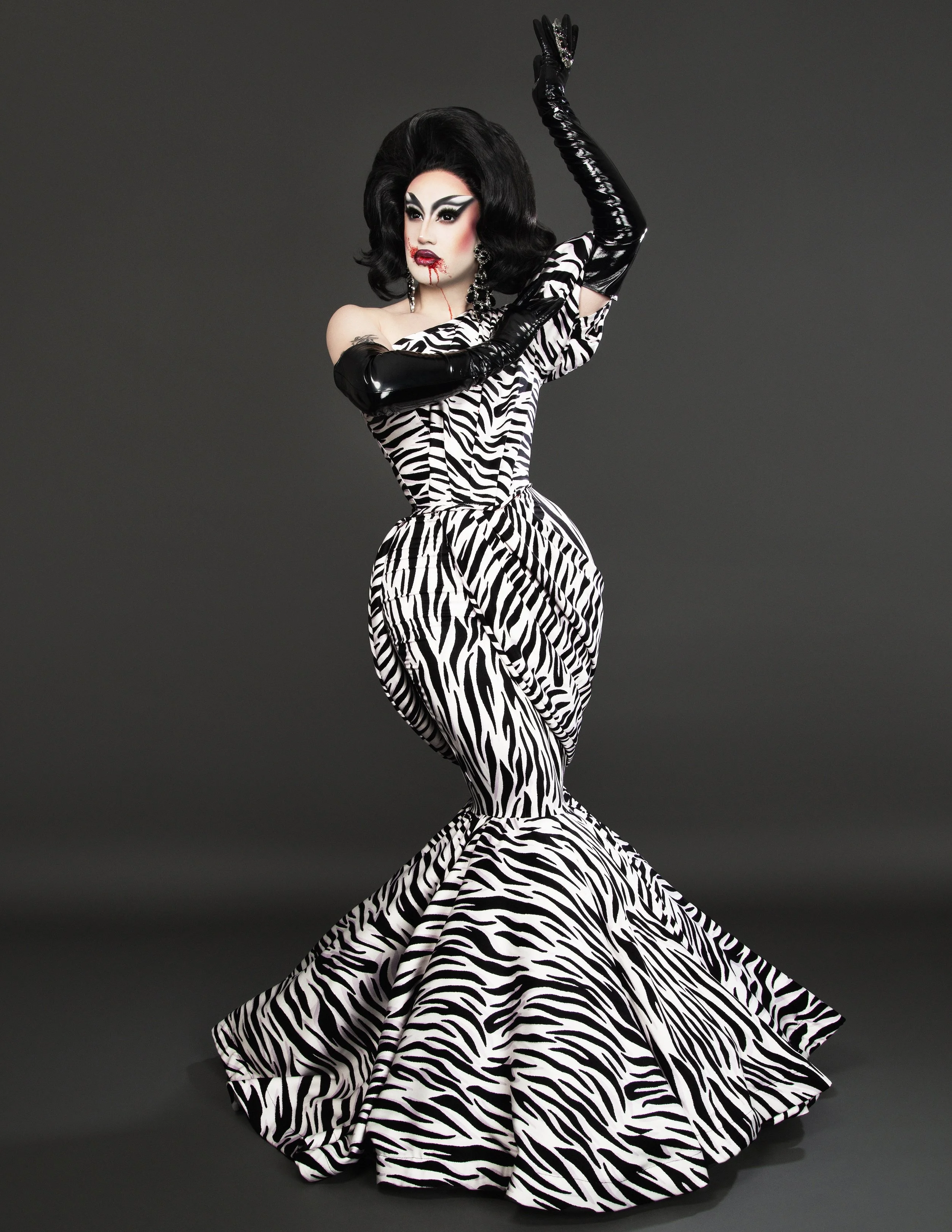 A person in a zebra-print dress with a matching train, black gloves, and bold makeup, posing against a dark background.