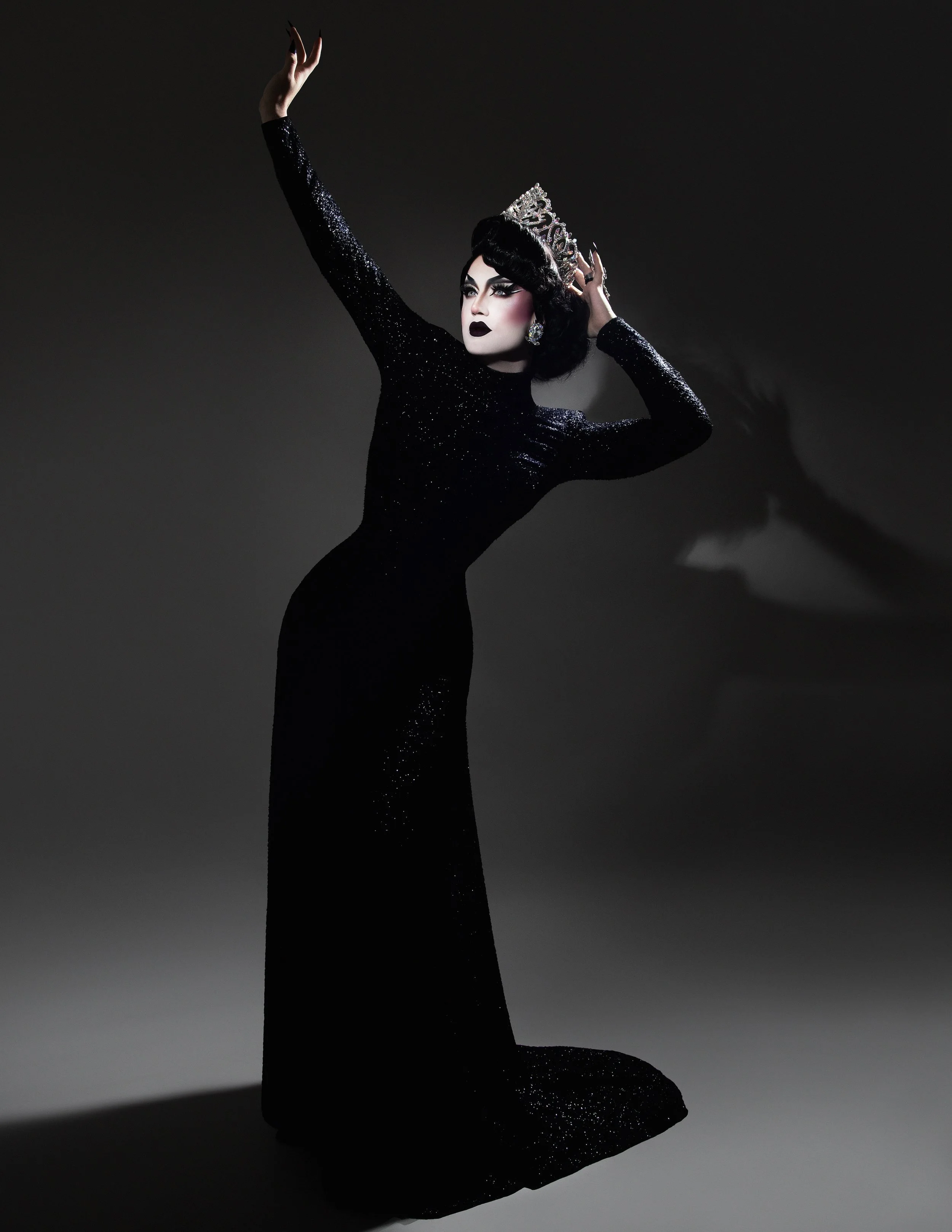A person dressed as a drag queen wearing a black, sequined, long-sleeved dress and a silver tiara, striking a dramatic pose with one arm raised and the other touching the tiara, set against a dark background.