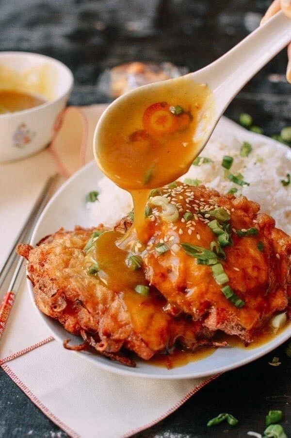 A spoon pouring gravy over a fried piece of chicken garnished with chopped green onions on a plate with rice.