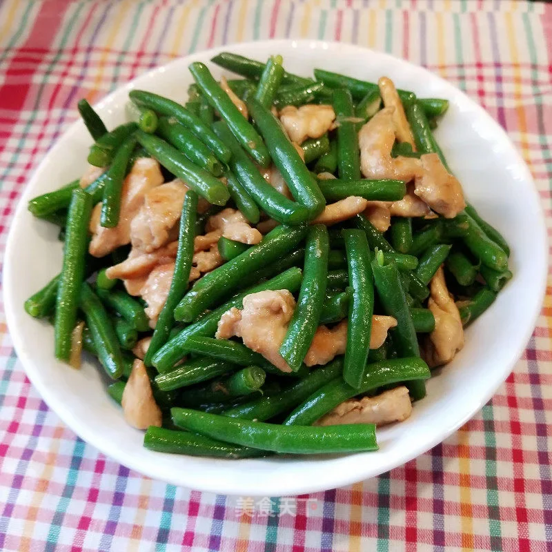 Stir-fried green beans with pieces of chicken in a white bowl on a checkered tablecloth.