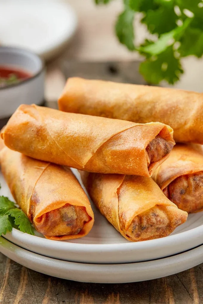 Six golden-brown spring rolls on a white plate with cilantro garnish, with a small bowl of dipping sauce in the background.
