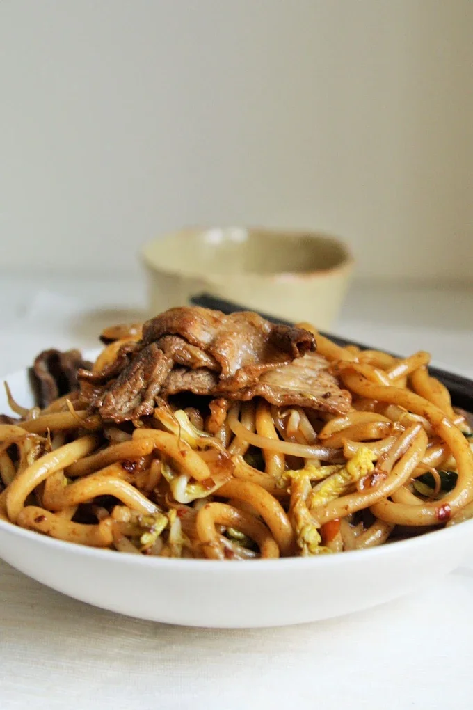 A bowl of stir-fried noodles topped with sliced beef and vegetables, with a small bowl of dipping sauce in the background.