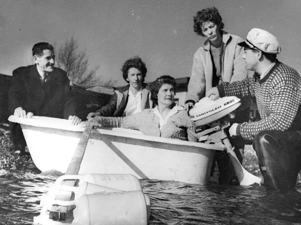 A group of five women and one man standing on a small boat in the water, with one woman sitting inside the boat. The man is operating an outboard motor, and they appear to be enjoying a boat outing outdoors.