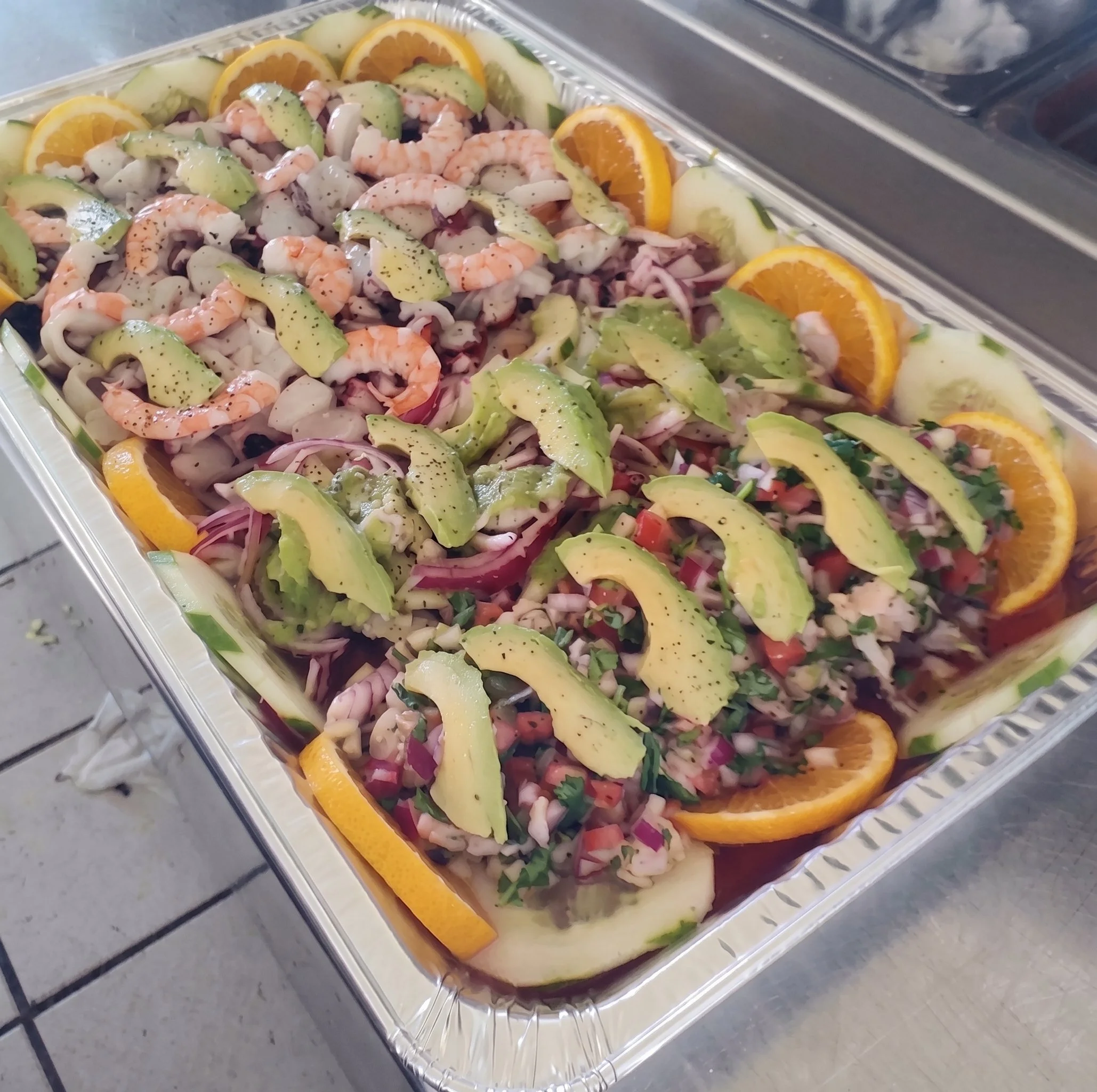 A seafood salad with shrimp, sliced avocado, red onions, chopped herbs, and orange slices garnished with black pepper, served in a clear plastic container.
