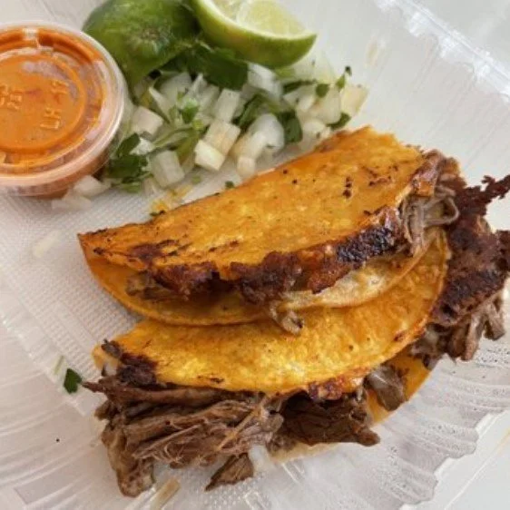 Close-up of a Mexican torta sandwich with shredded beef, topped with corn, served with a side of chopped onions, cilantro, lime, and hot sauce.