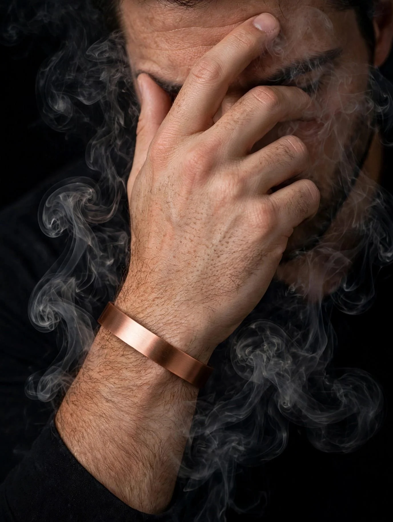 Close-up of a man with light skin and facial hair, holding his face with one hand, with smoke swirling around him, wearing a copper-colored bracelet on his wrist.