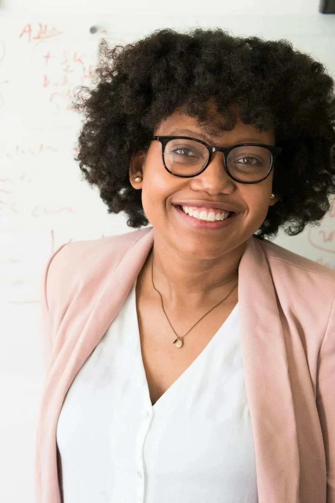 Smiling woman with curly hair and glasses, wearing a pink blazer over a white top, with a gold heart pendant necklace, standing in front of a whiteboard with red writing.