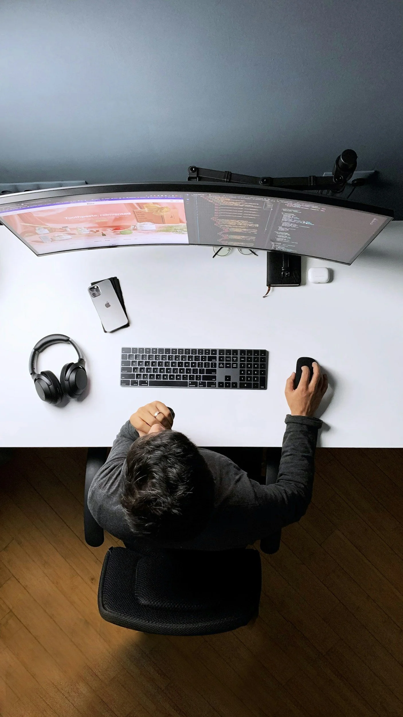 Top-down view of a person sitting at a desk with a curved monitor, keyboard, wireless mouse, headphones, and an iPhone, working on a computer with code visible on the screen.