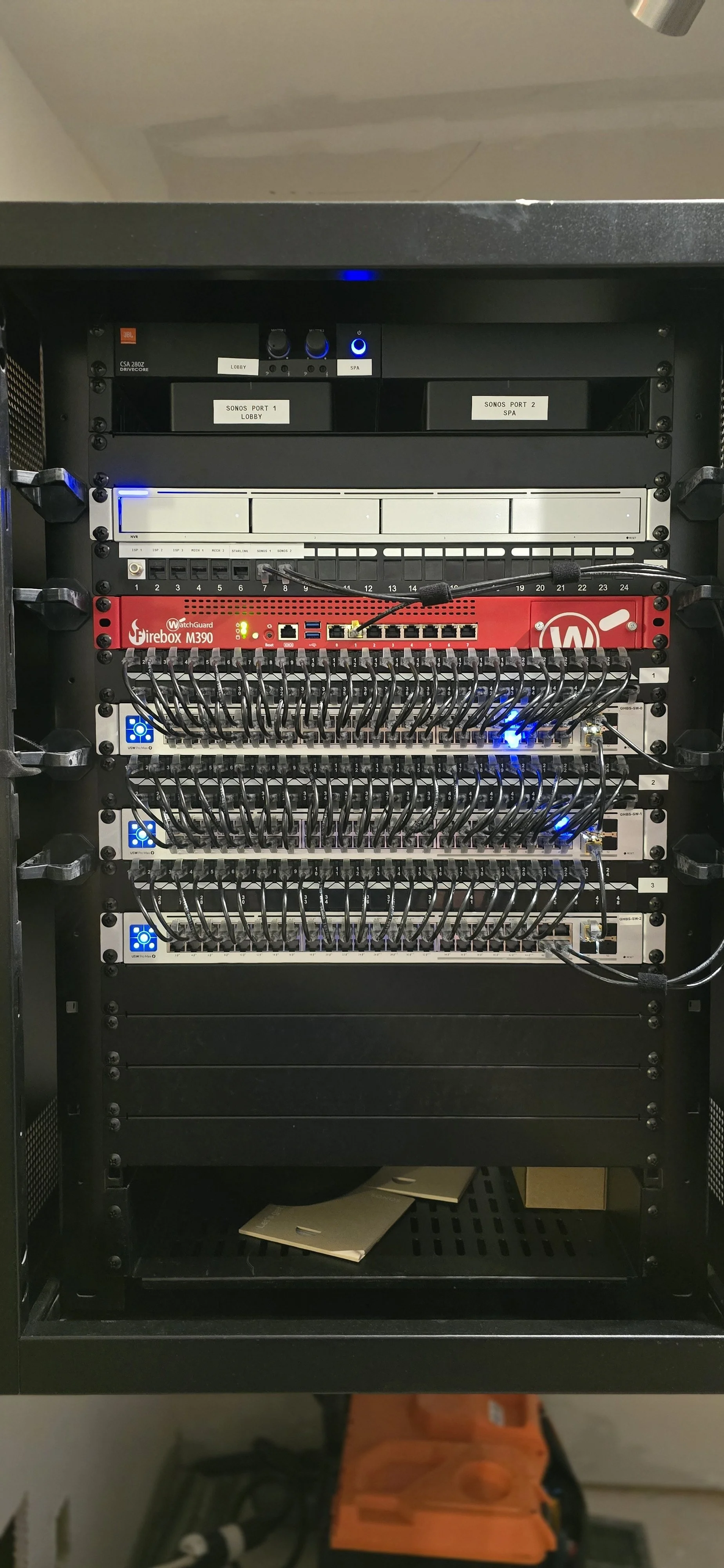 A server rack with multiple network switches and hardware components, including a Firebox M390 device in red, all interconnected with numerous black cables.