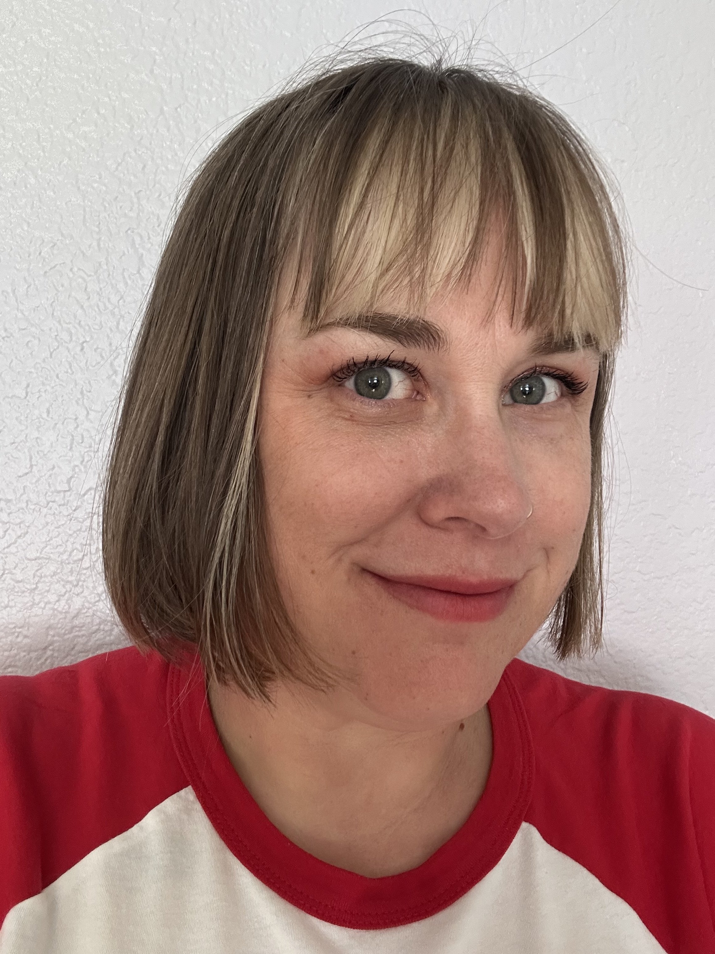 A woman with light brown hair in a bob cut, wearing a red and white baseball-style shirt, smiling slightly, standing against a white textured wall.