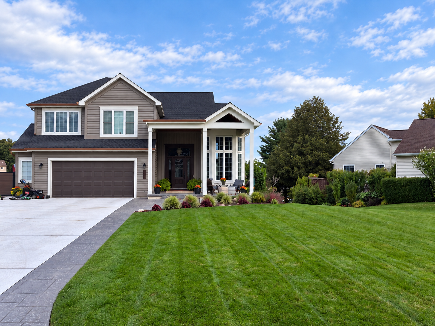 Front view of a two-story house with a manicured lawn, a concrete driveway, large windows, and a cozy porch with seating, surrounded by trees and neighboring homes under a partly cloudy sky.