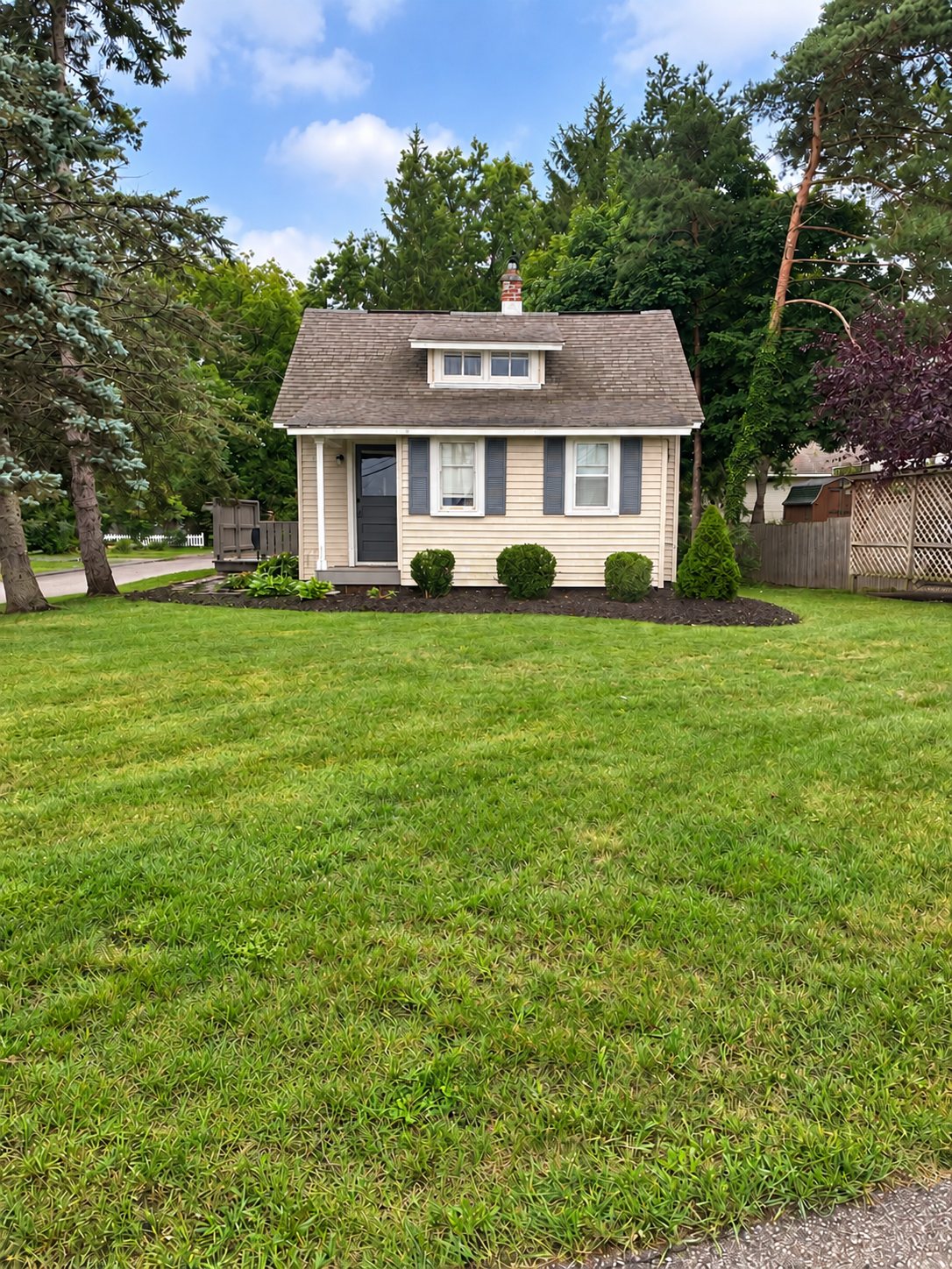 Small beige house with dark shutters and a small front porch, surrounded by a neatly maintained lawn and bushes, with tall trees in the background under a partly cloudy blue sky.
