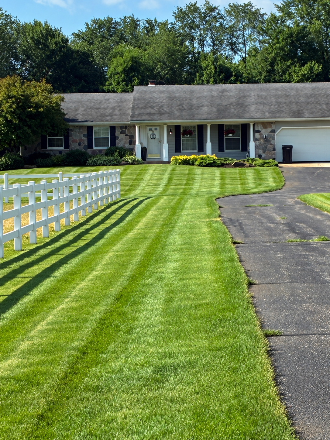 Front yard with freshly mowed green grass, a white picket fence, a paved driveway, and a house with a stone exterior, black shutters, and hanging flower baskets.
