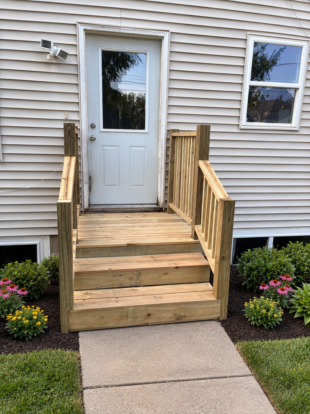 New wooden ramp with railings leading to a white door on the exterior of a house, with flowers and shrubs in the front yard.