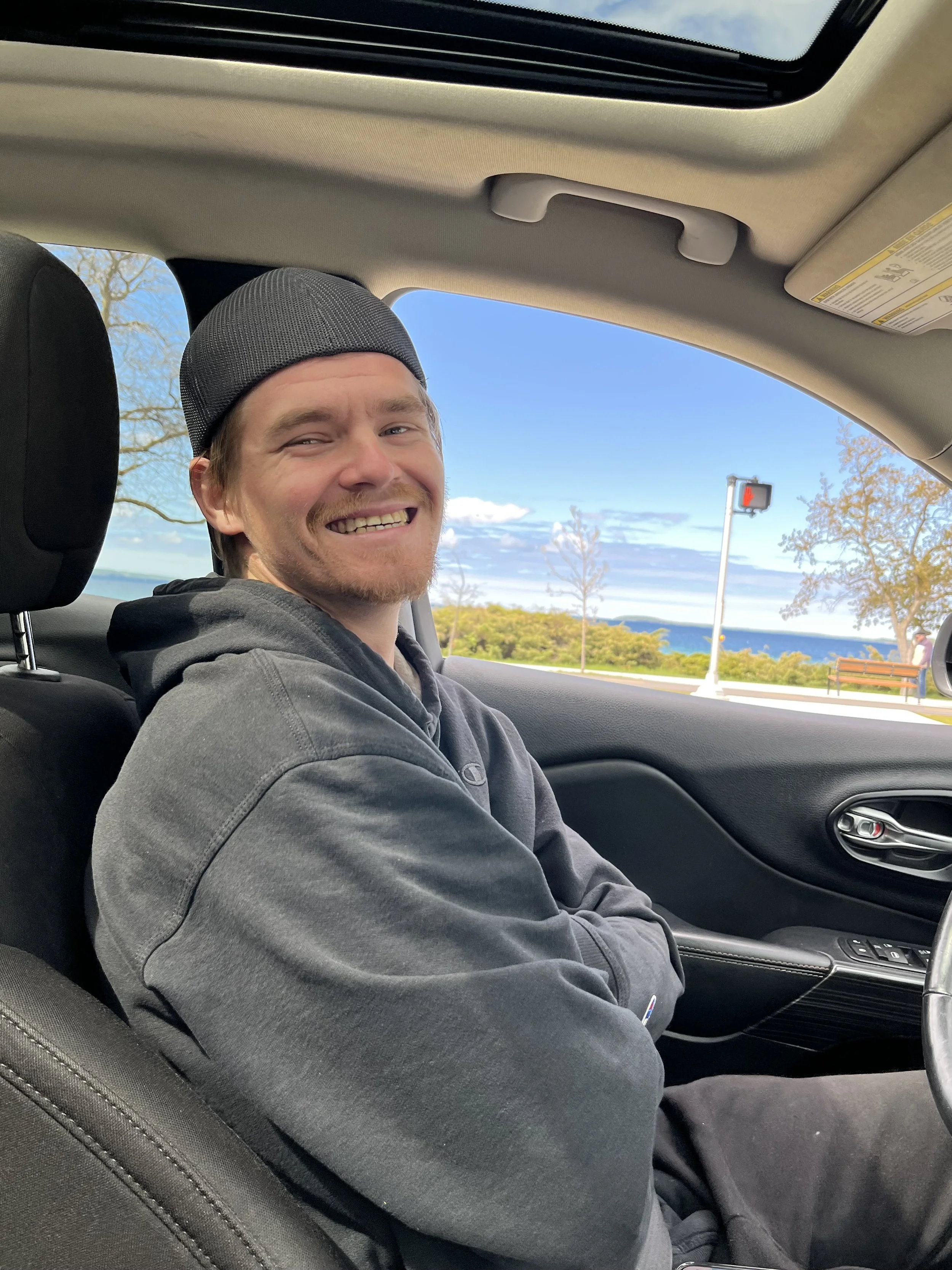 A smiling man wearing a dark hoodie and a gray baseball cap sitting in the passenger seat of a car, with scenic outdoor landscape visible through the window including trees, a bench, and a body of water under a partly cloudy sky.
