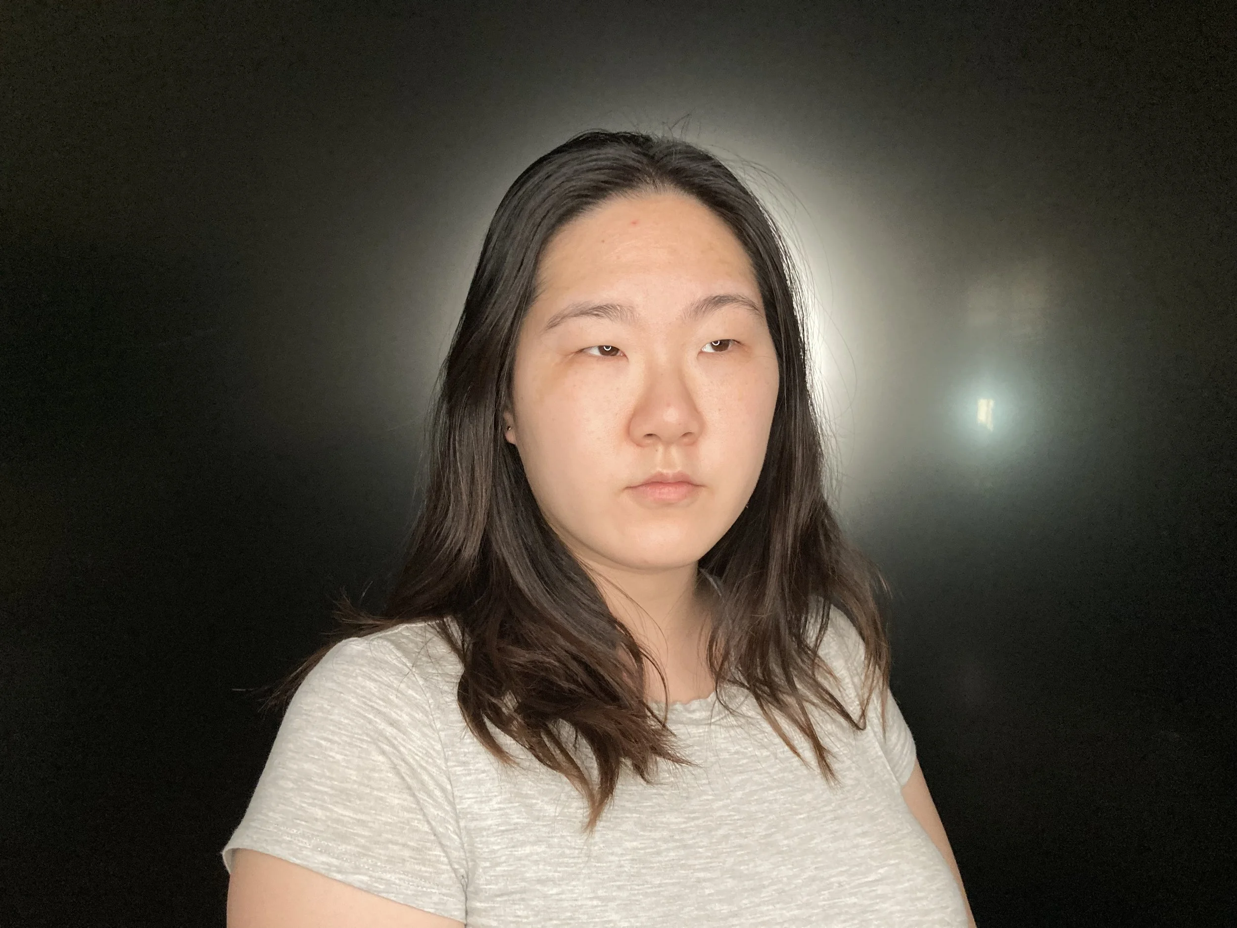 A woman with long dark hair wearing a light gray shirt standing in front of a dark background with a halo-like light behind her head.