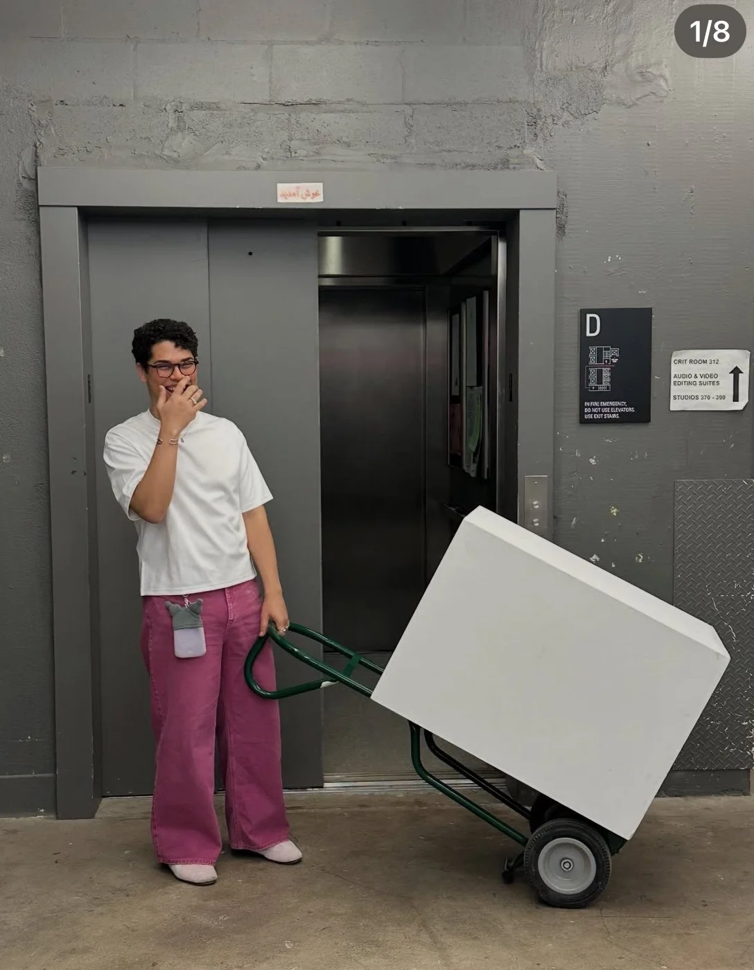 Person in white shirt and pink pants standing next to a hand truck loaded with a large white box, in front of an elevator in a gray industrial building.