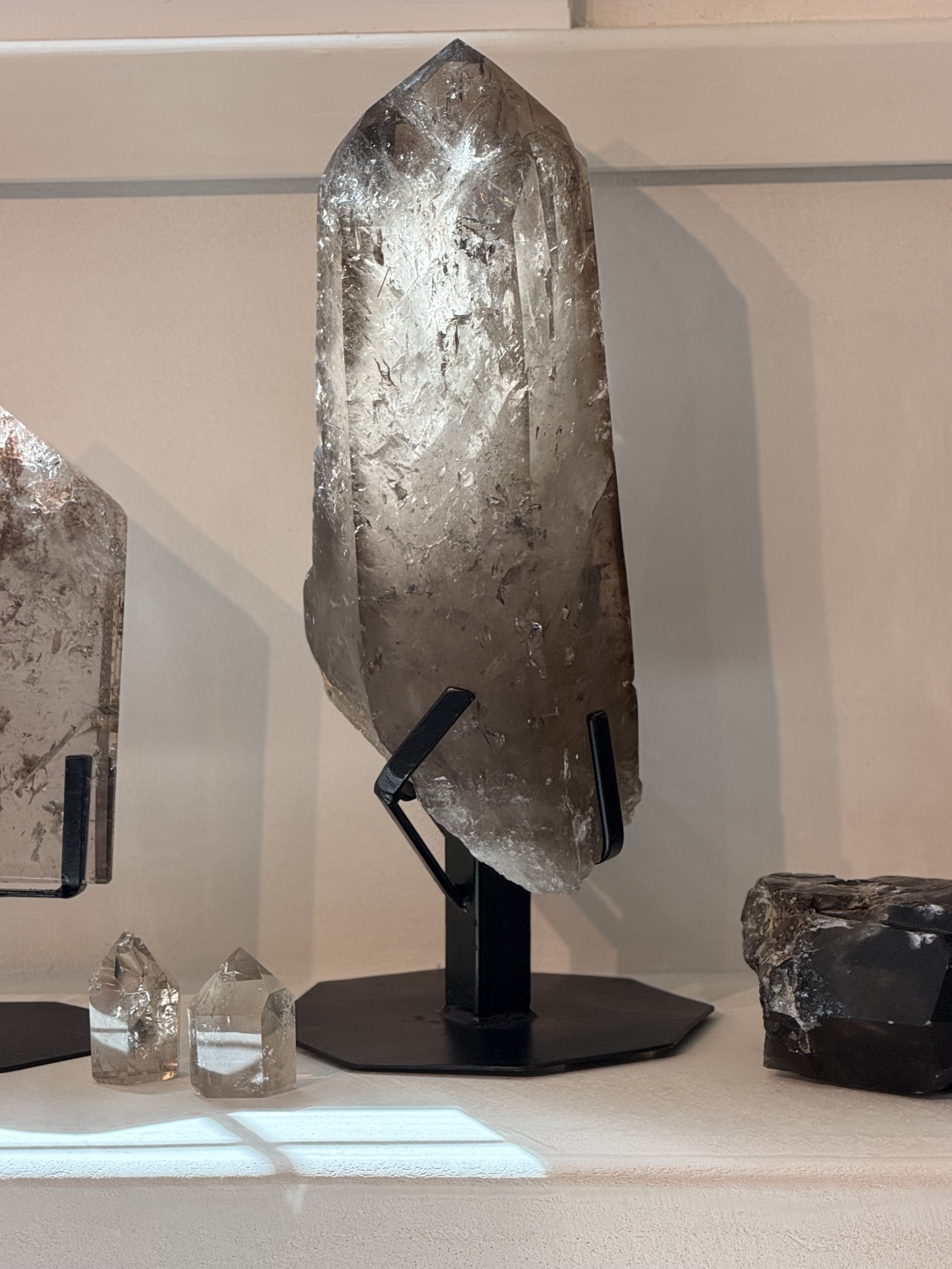 A large, clear, pointed crystal displayed on a black stand, with smaller crystals nearby on a white surface.