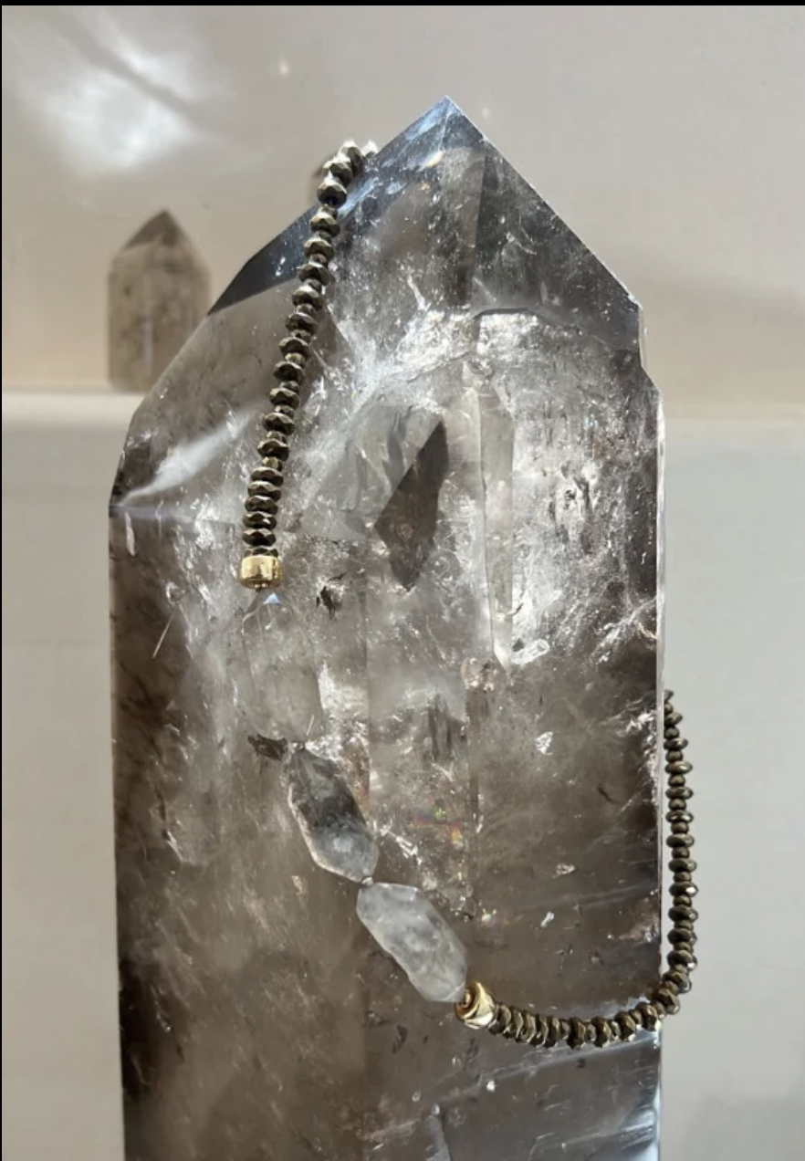 A large, clear quartz crystal with a necklace draped over its top, featuring small dark beads and a few larger clear stones.