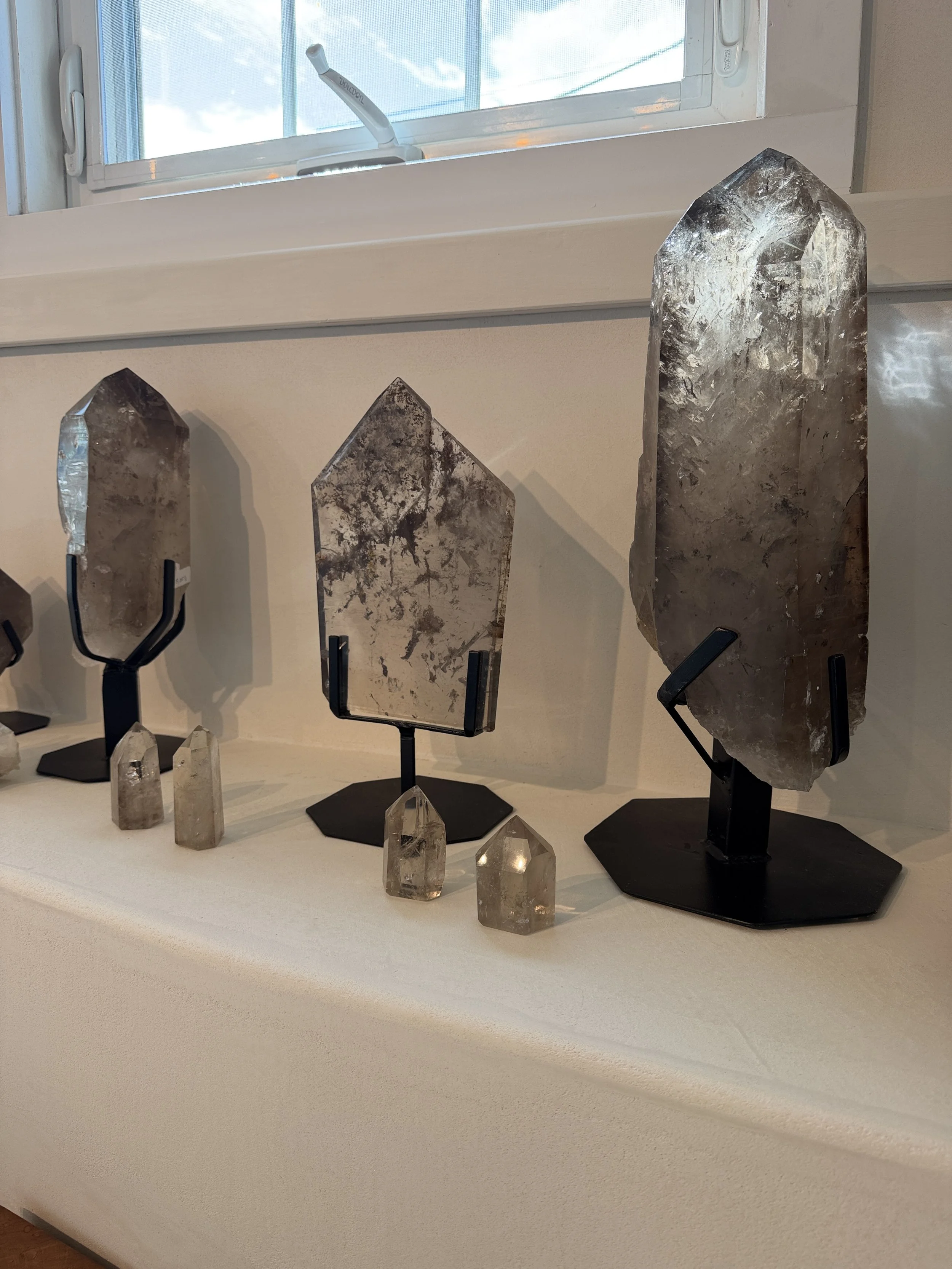 Display of multiple polished crystals on black stands and smaller raw crystals on a white surface near a window.