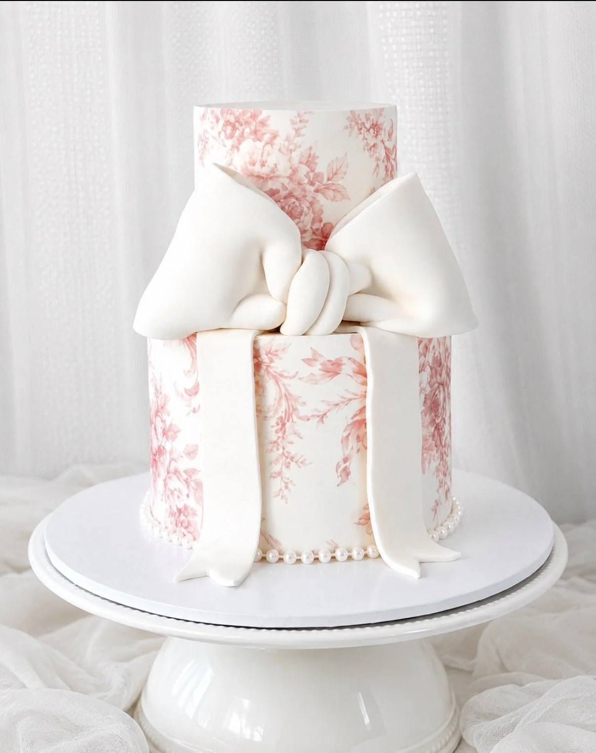 Elegant cake decorated with a pink floral pattern, large white bow, pearls, and draped fondant, on a white cake stand.