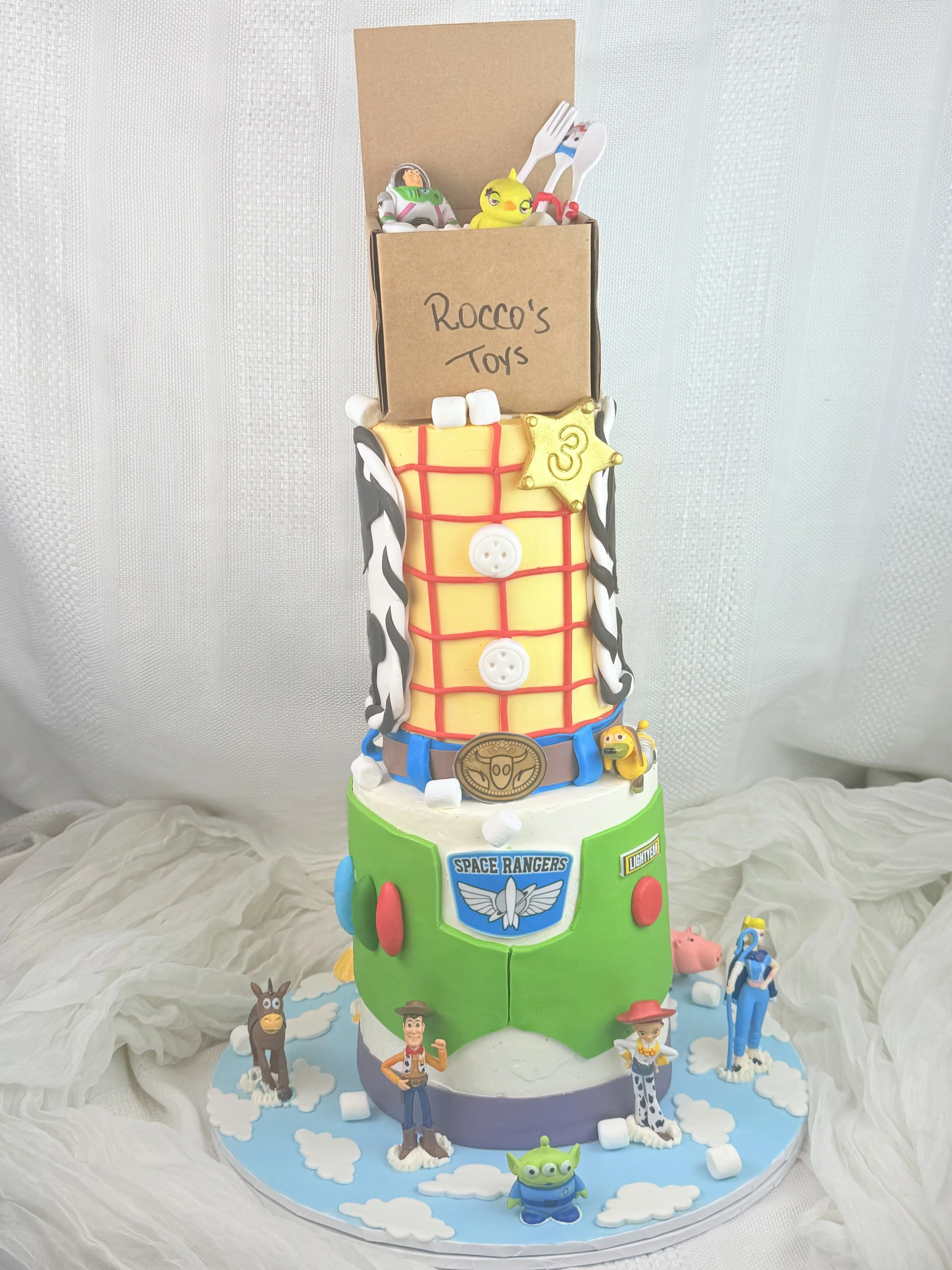 A Toy Story themed birthday cake featuring characters from the movie, including Woody, Buzz Lightyear, and other toys, with a colorful, tiered design resembling a toy box and space ranger logo.