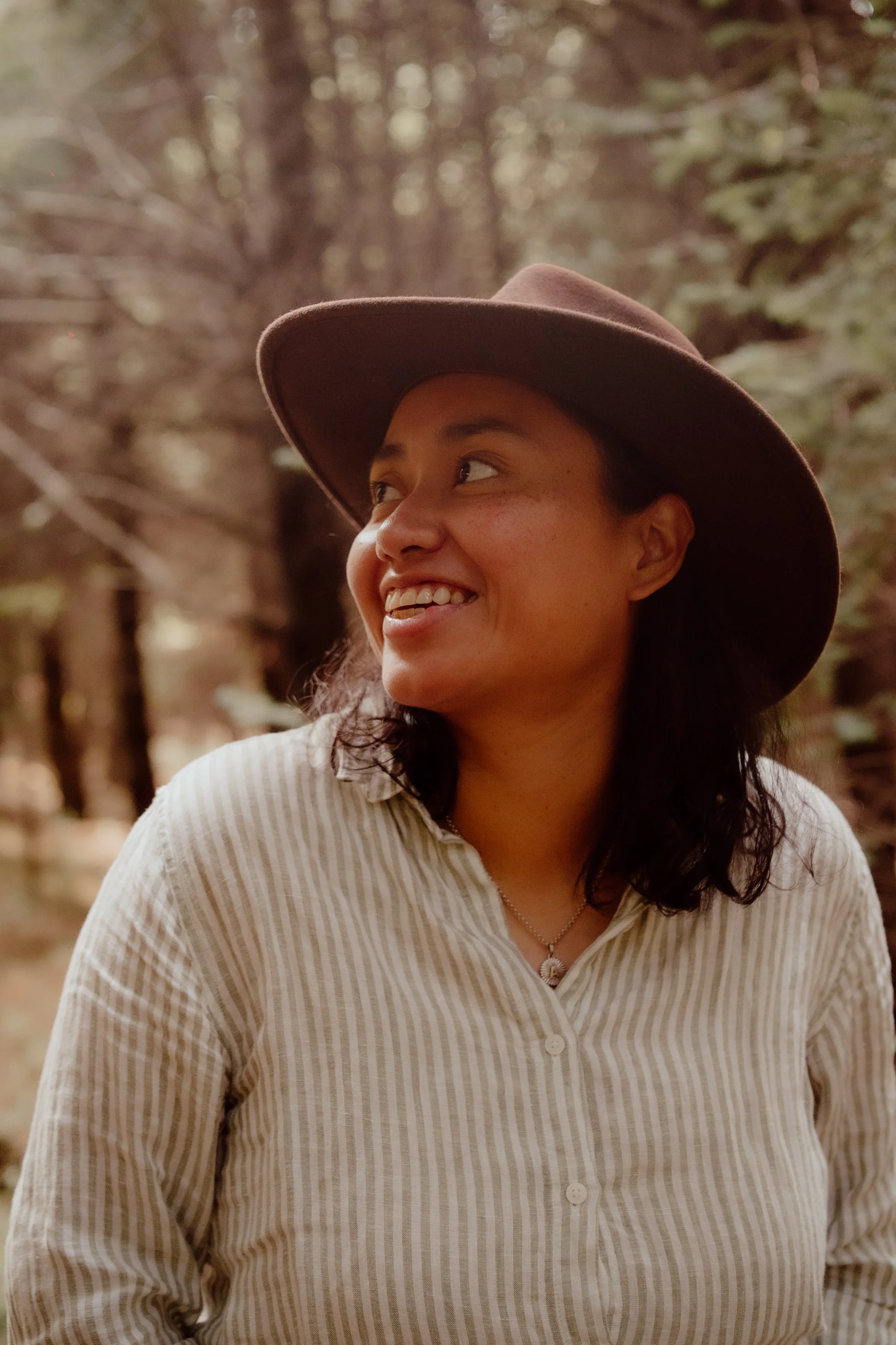 Mujer sonriendo, lleva sombrero y camisa de rayas, en un entorno natural con árboles.