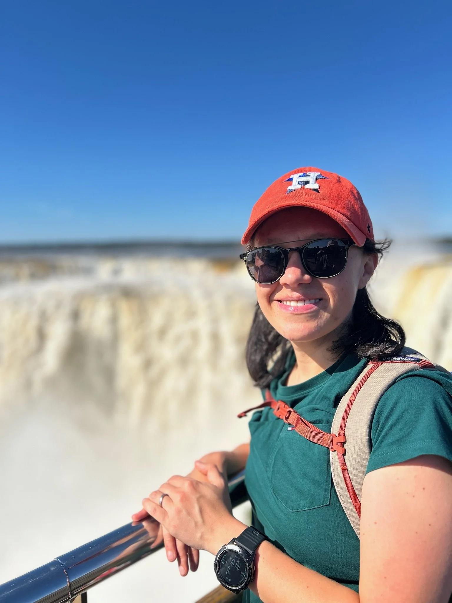 Mujer con gafas de sol, gorra roja y reloj, sonriendo, en un mirador con vista a un paisaje de agua y cielo azul.