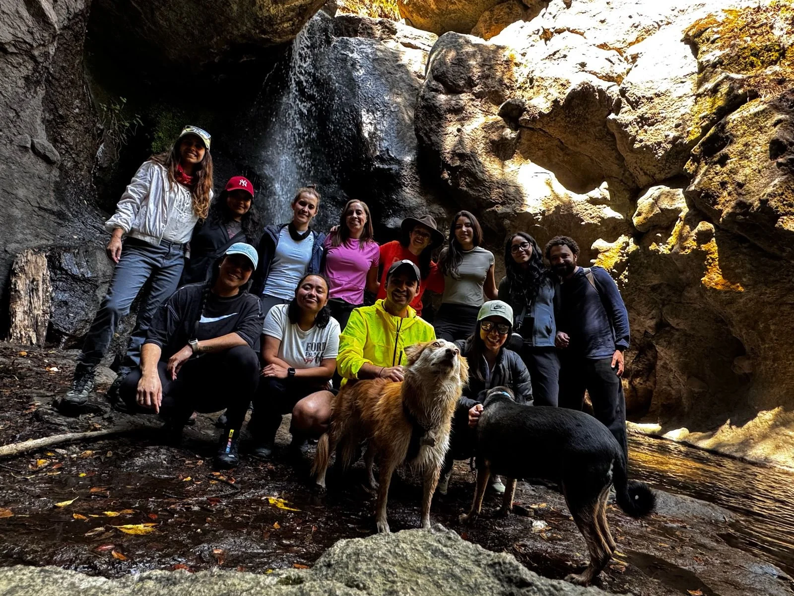 Grupo de senderistas posando sonrientes junto a sus perros frente a una cascada y pared rocosa durante una caminata de hiking pet friendly en la naturaleza.
