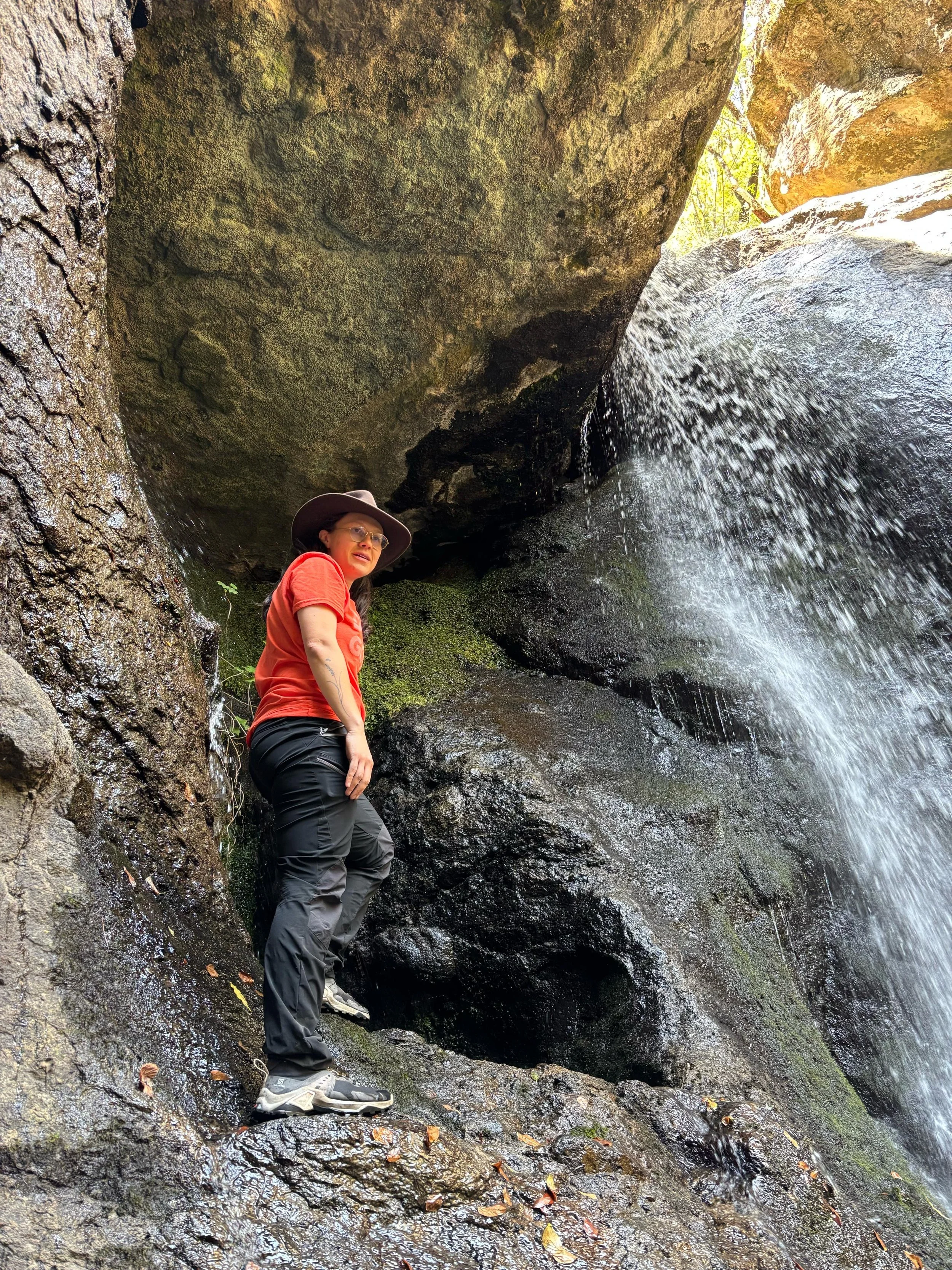 Una mujer con sombrero, gafas, camiseta naranja y pantalones oscuros de pie en una roca húmeda en un cañón con agua cayendo de una cascada pequeña a su lado.