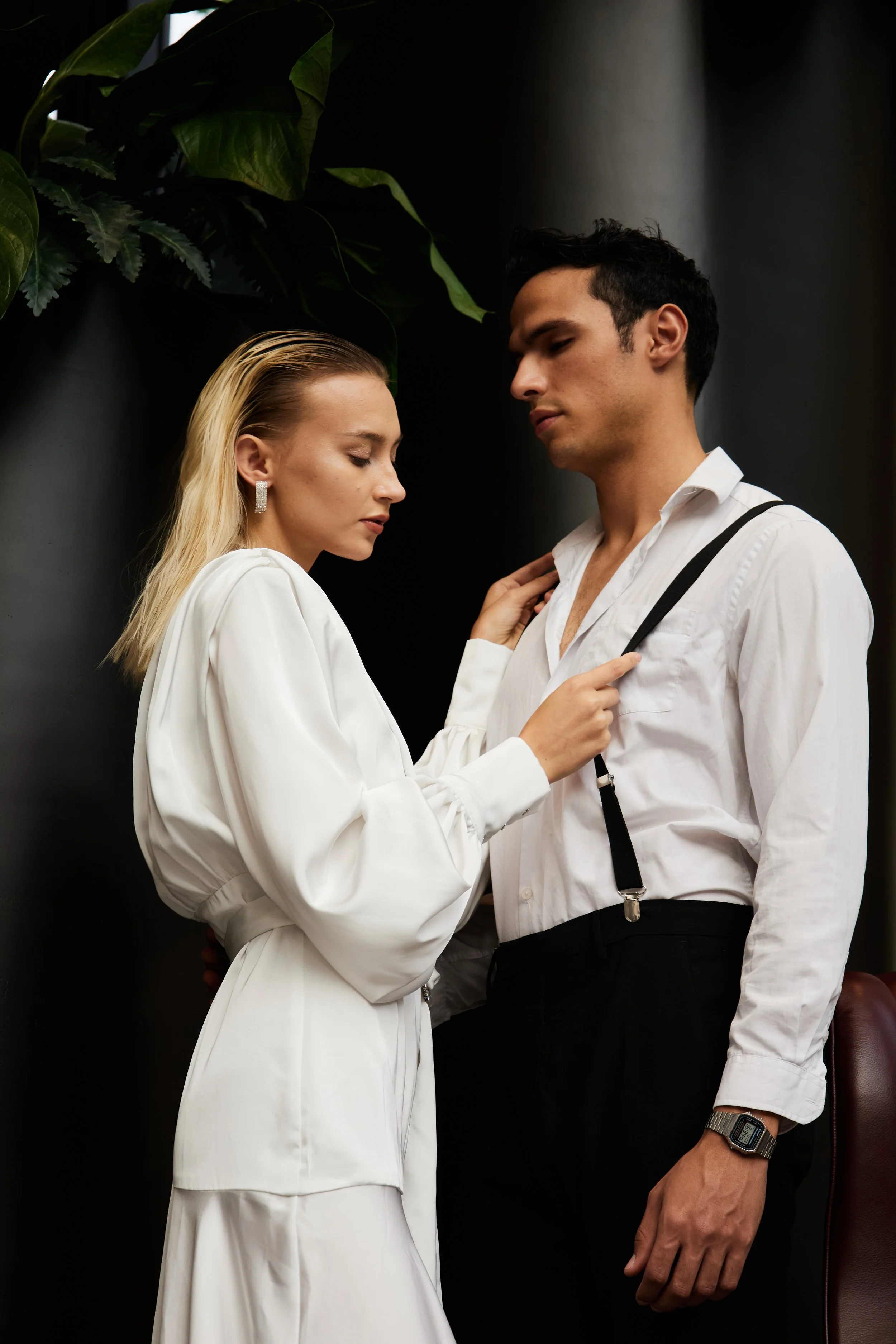 A woman in a white dress with blonde hair and earrings is gently touching the chest of a man in a white shirt with suspenders, who is wearing a watch and has dark hair, against a dark background with large green leaves.