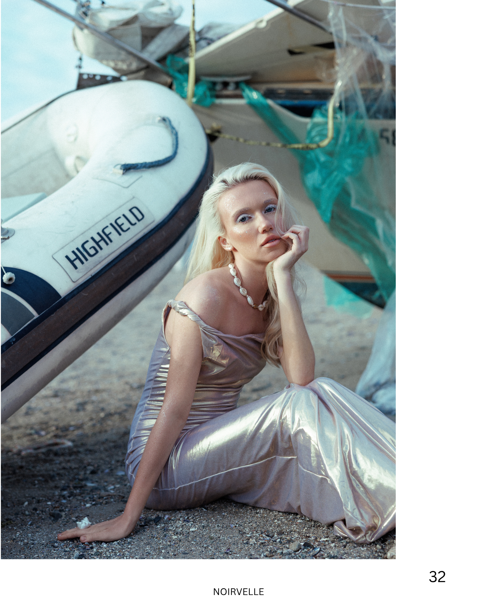 A woman with platinum blonde hair and metallic makeup poses next to a small boat labeled 'Highfield' on a beach. She is dressed in iridescent clothing and jewelry, resting her chin on her hand with her elbow on her knee, looking at the camera.