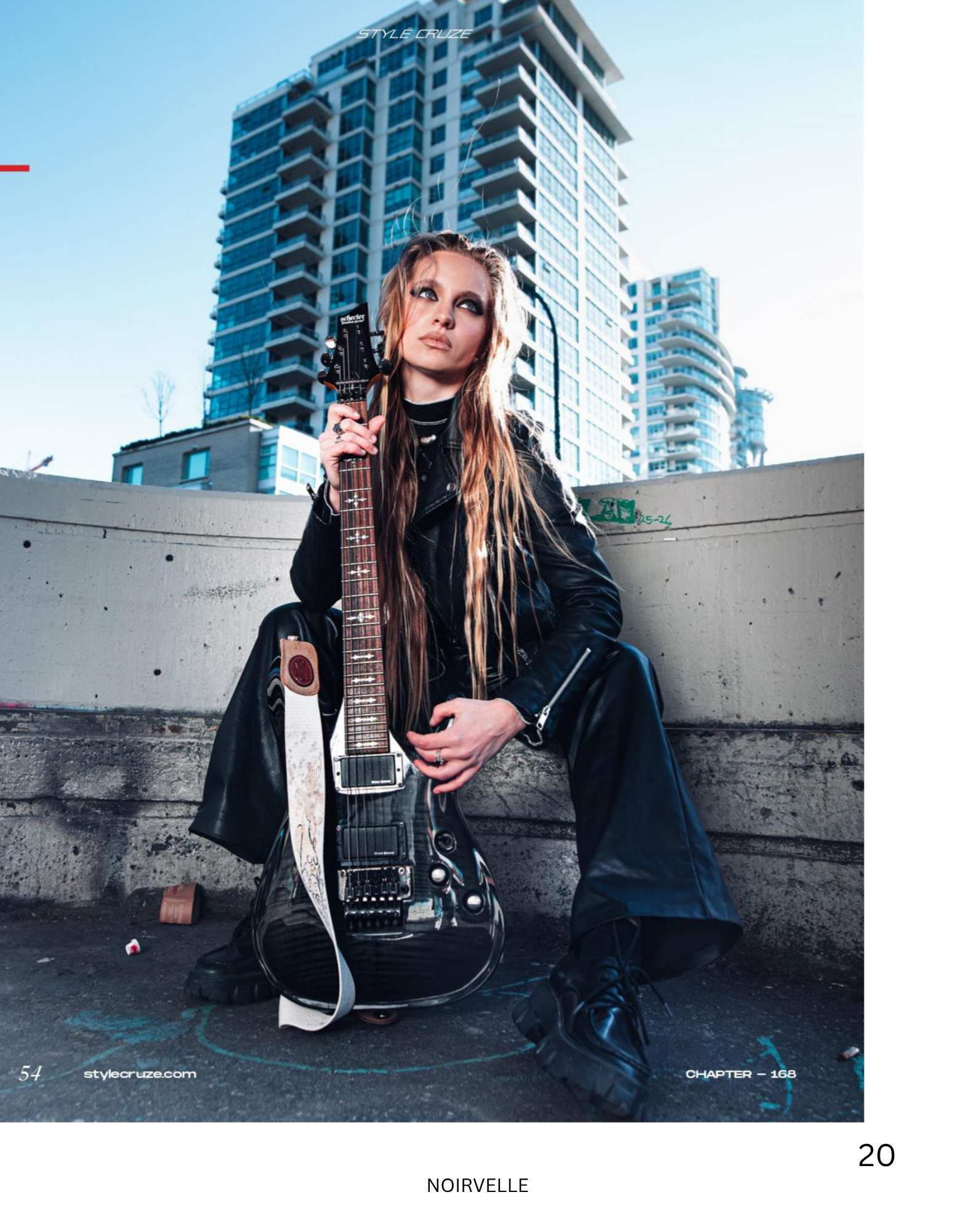 A woman with long, wavy brown hair and light makeup sitting outdoors with a bass guitar resting against her left shoulder. She is wearing a black leather jacket, black pants, and black boots. Behind her are modern high-rise apartment buildings, and s