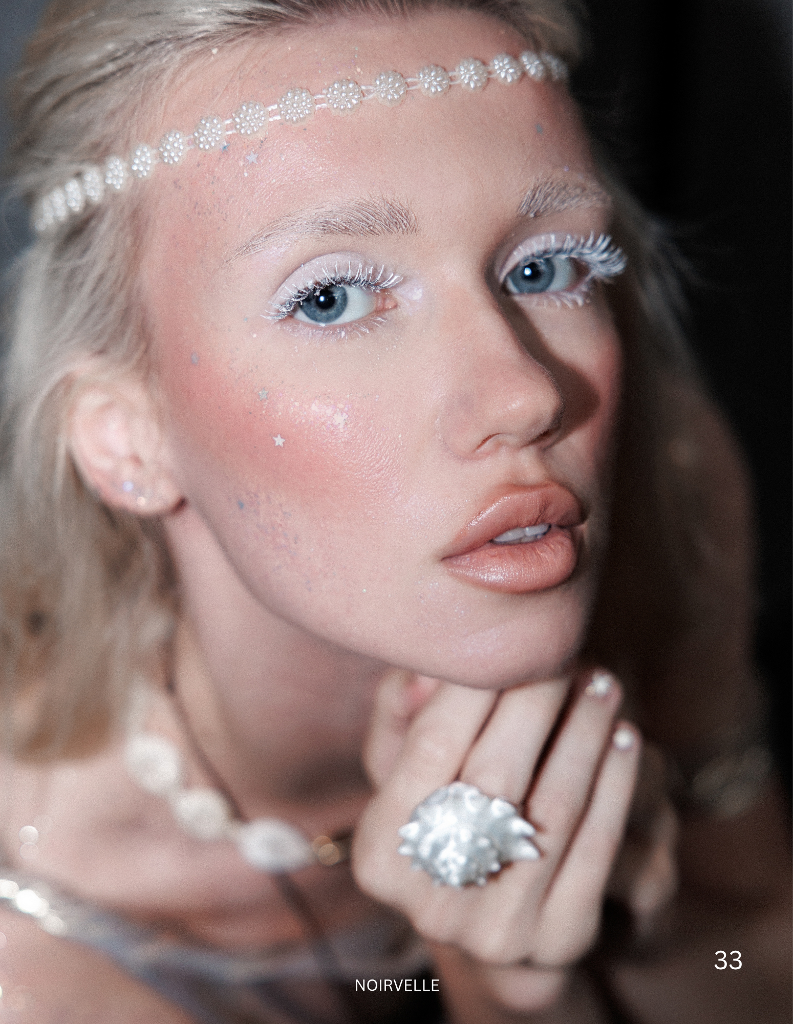 Close-up of a woman's face with platinum blonde hair, wearing a beaded headband, with glittery makeup, light blue eyes, and star-shaped glitter on her cheeks, adorned with a large silver ring and bracelet.
