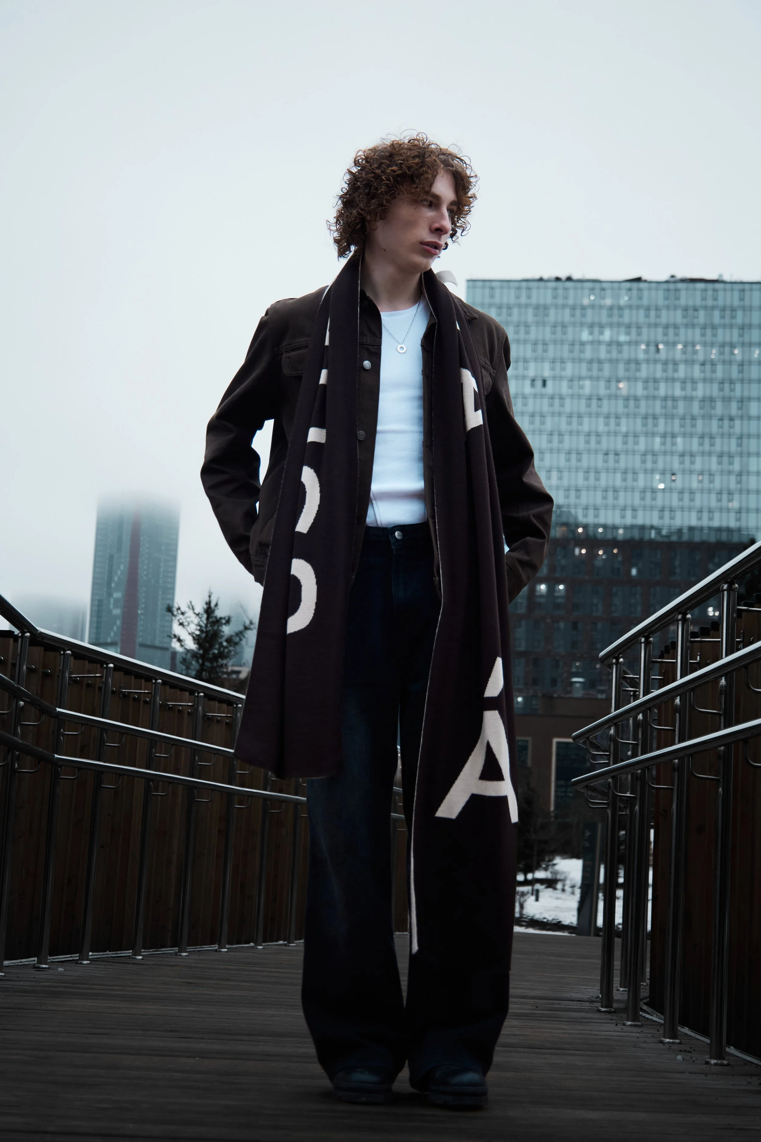 Person with curly hair wearing a dark coat and a scarf, walking outdoors on a wooden bridge with modern buildings and a cloudy sky in the background.