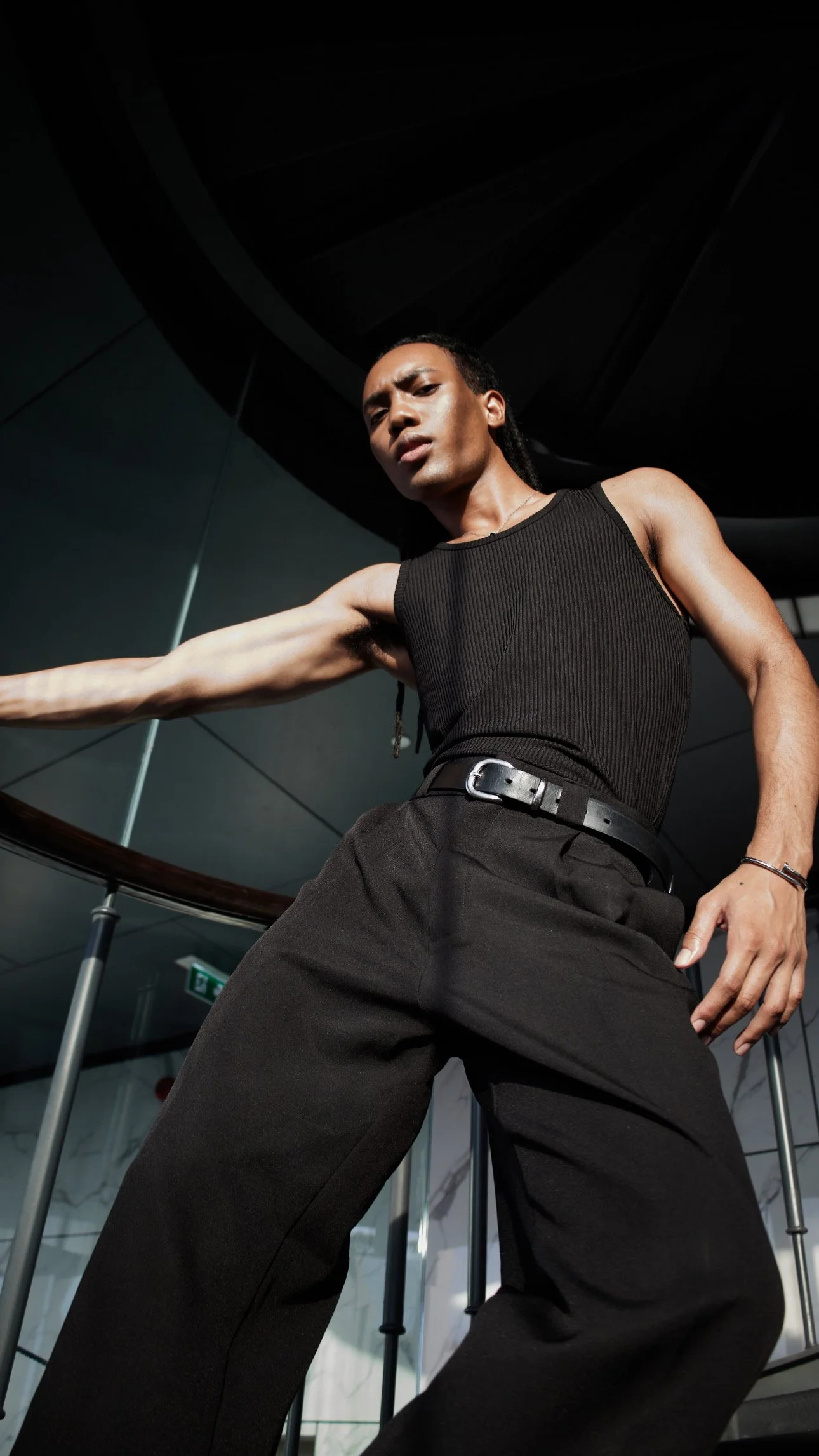 A person with dark skin and long braided hair wearing a black ribbed tank top and black pants, striking a confident pose in an interior space with a spiral staircase and glass walls.