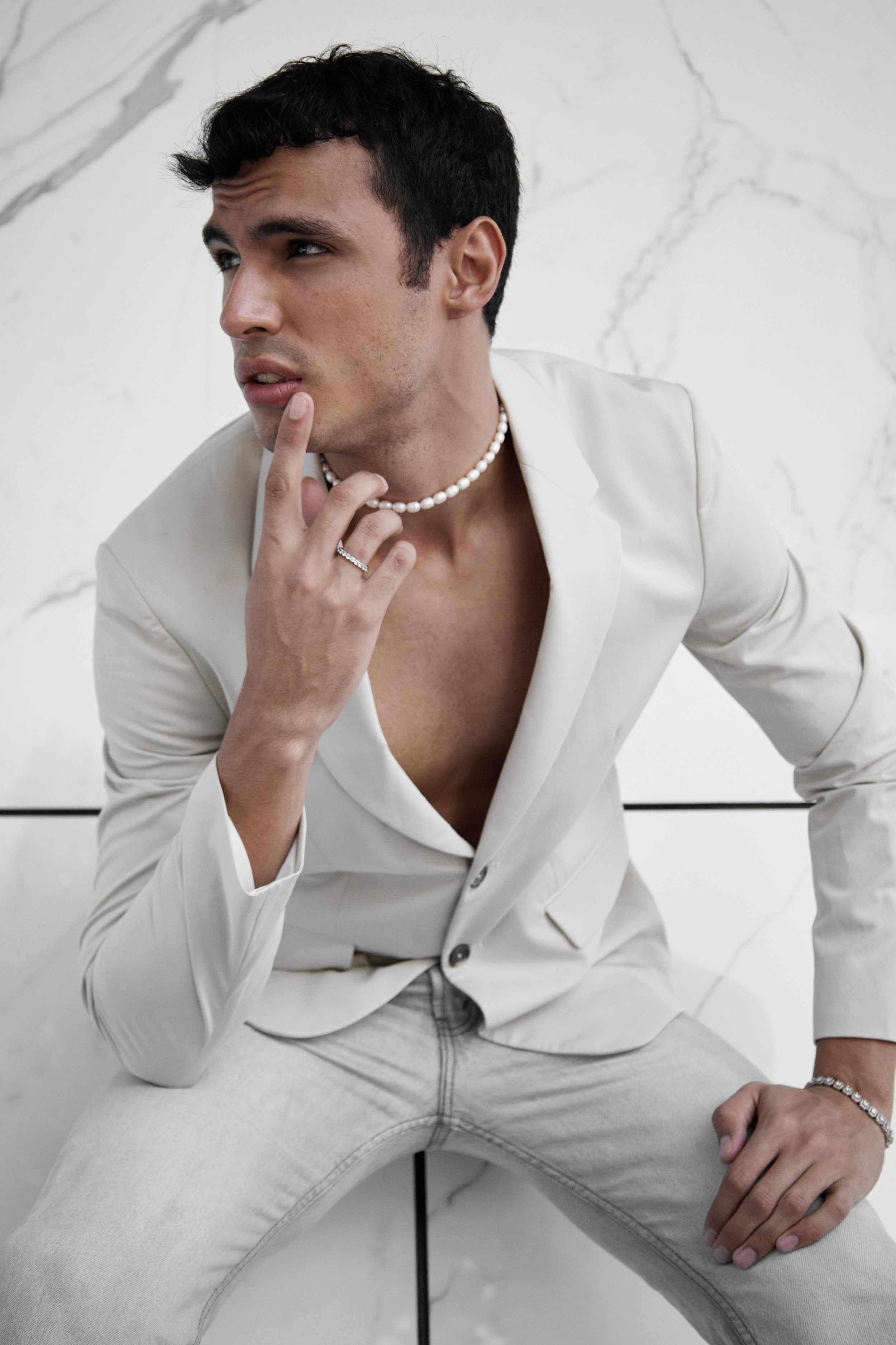 A young man with black hair, wearing a white blazer, pearl necklace, and rings, sitting with one leg crossed over the other, touching his chin thoughtfully, against a white marble background.