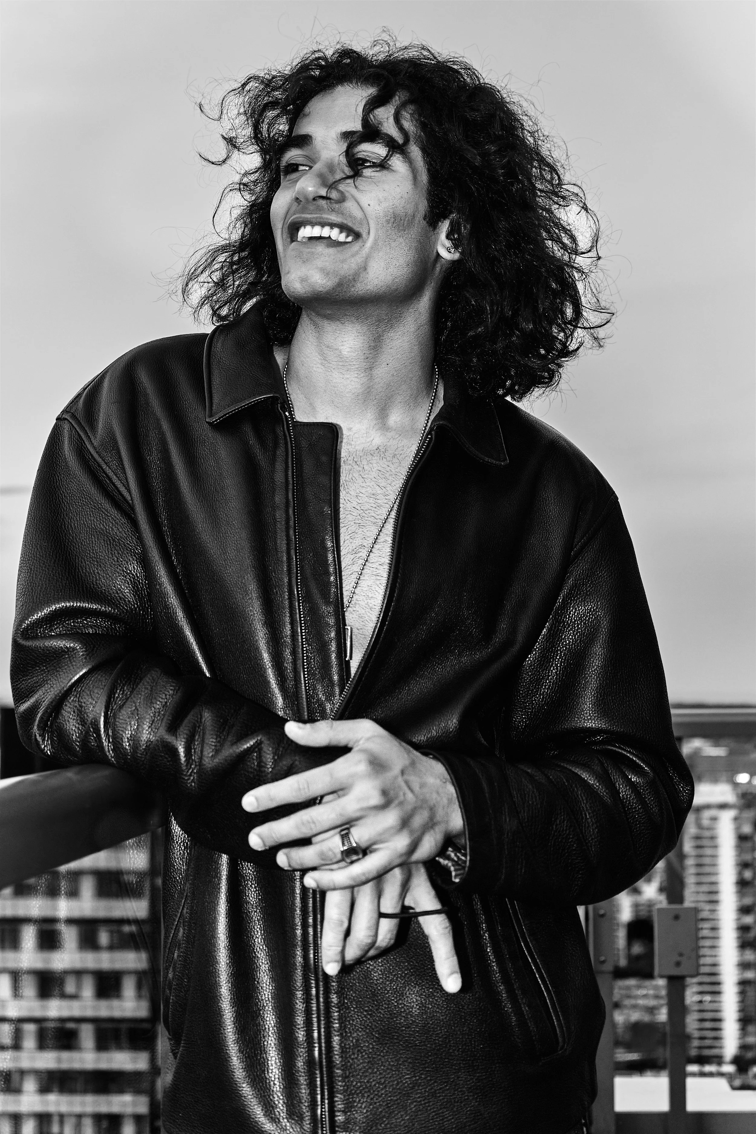 A person with curly hair leaning on a railing with a cityscape in the background, smiling and looking to the side, in black and white.