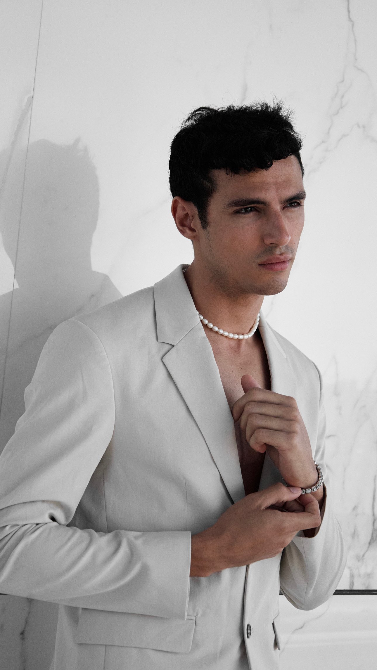 A young man with dark curly hair and fair skin is dressed in an elegant white suit and jewelry, including a pearl necklace and bracelet. He has a serious expression and is standing in front of a white marble wall with subtle gray veining.