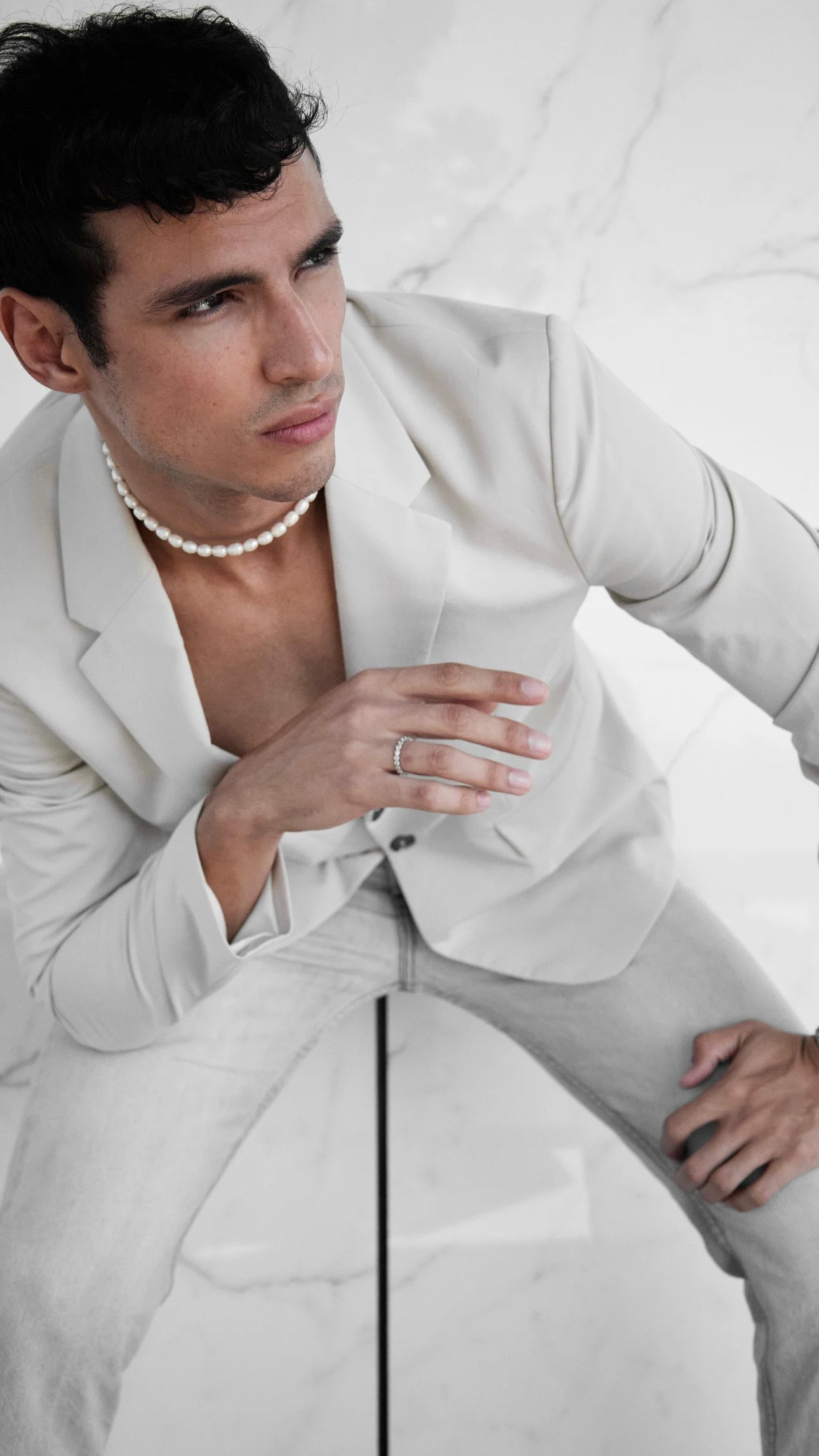 A young man with dark hair, wearing a white suit and pearl necklace, is sitting on a marble floor, looking thoughtfully to the side, with one hand resting on his knee.
