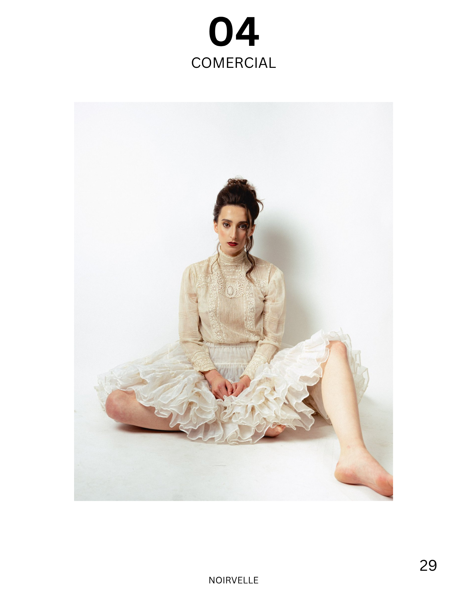 A woman with dark hair styled in a bun, wearing vintage cream-colored lace blouse and a ruffled cream skirt, sitting on the floor against a plain white background.