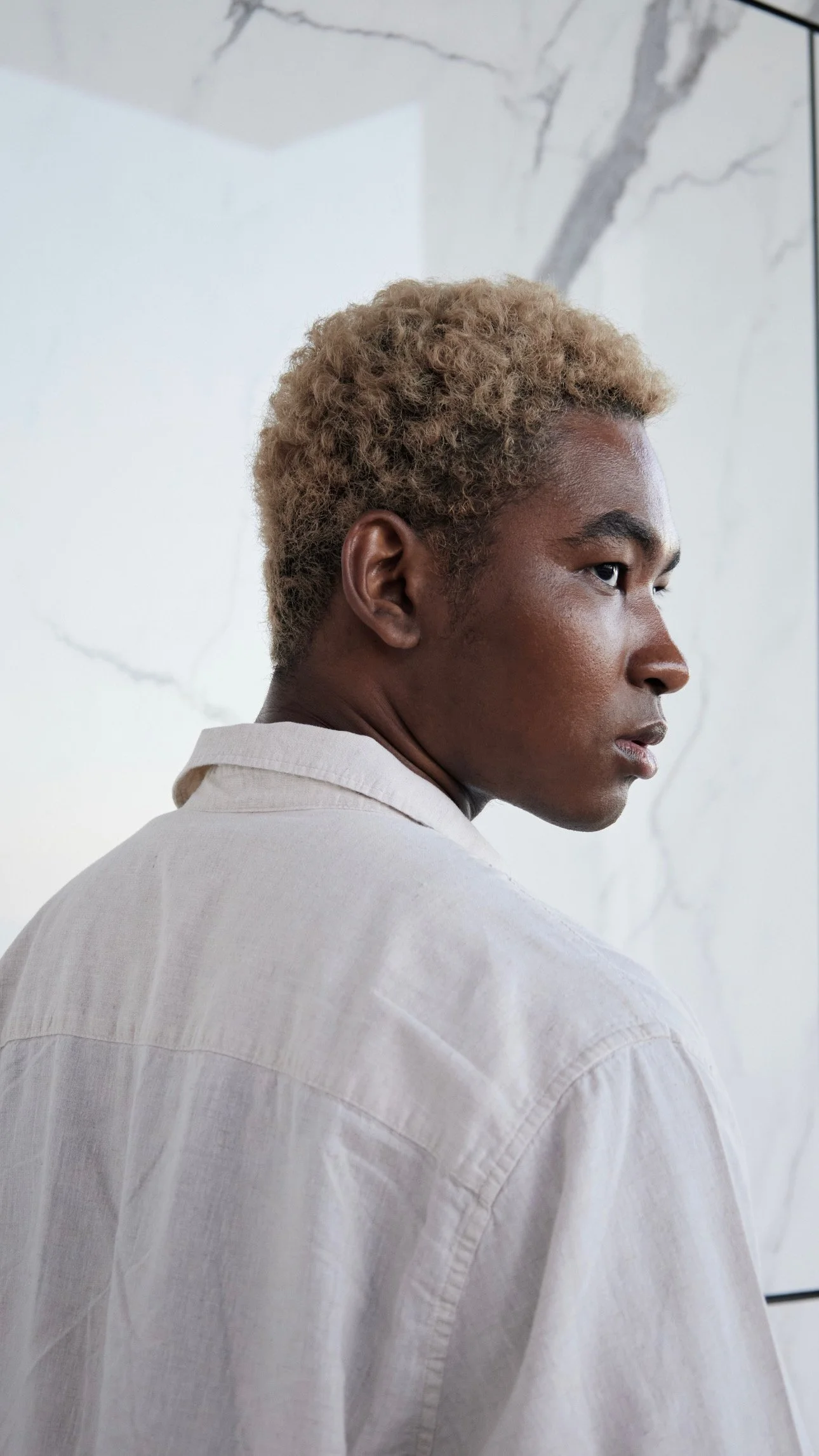 Side profile of a person with short, curly blonde hair wearing a light-colored shirt in front of a marble wall.