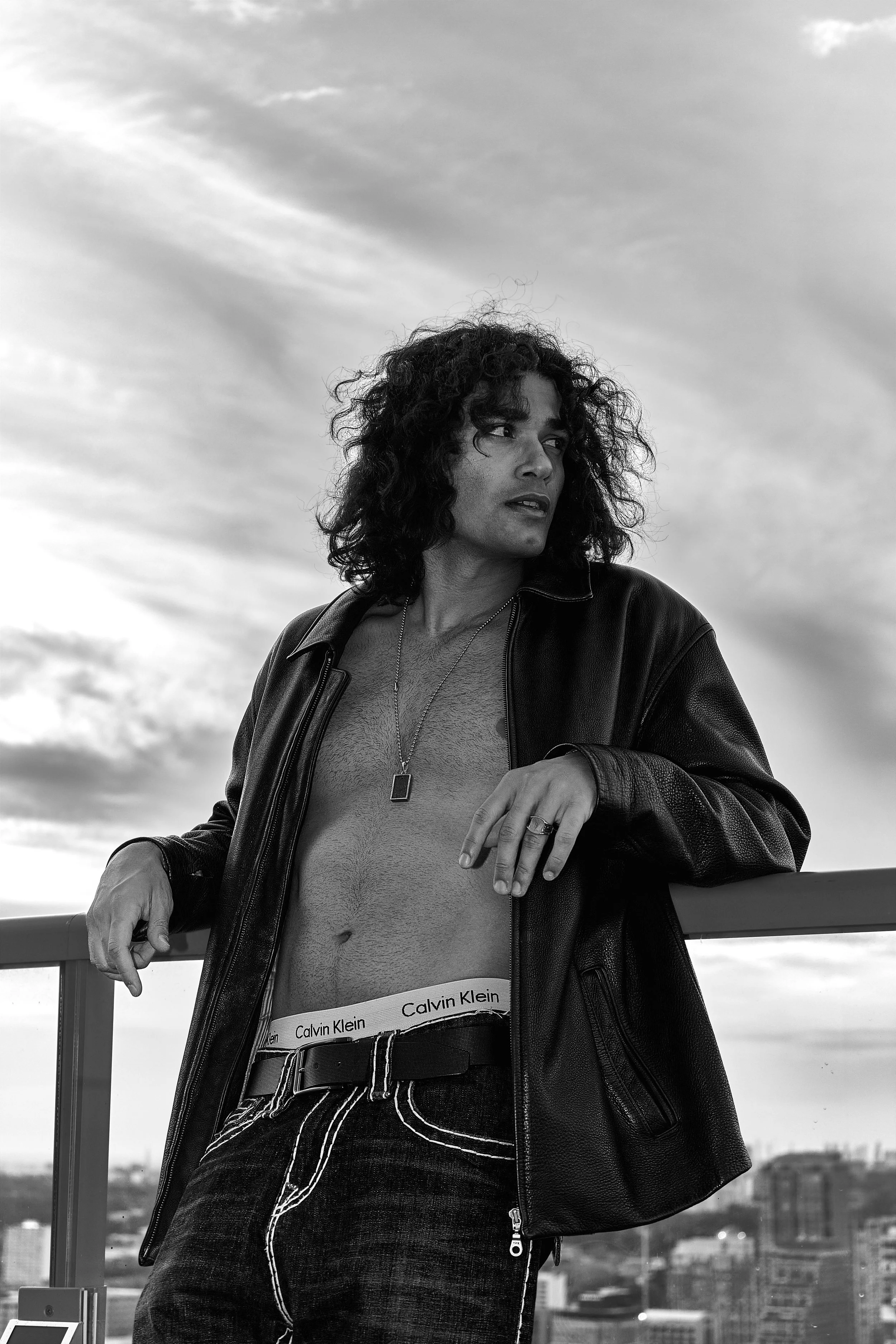 Black and white photo of a woman with curly hair wearing a leather jacket, Calvin Klein underwear, and striped jeans, leaning on a railing with a cityscape in the background.