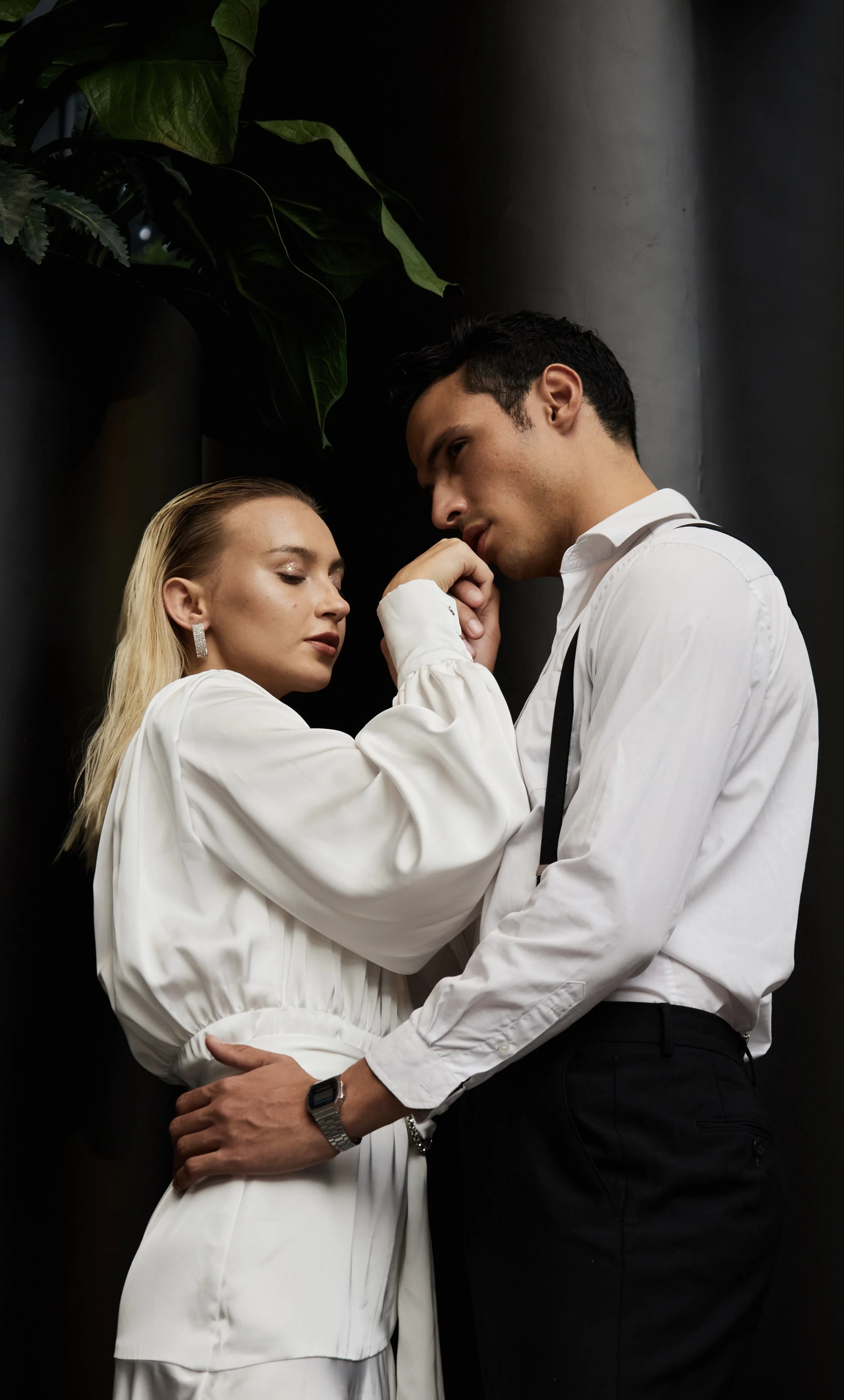A man and woman in formal attire are close together, touching foreheads and gazing into each other's eyes, against a dark background with green foliage.